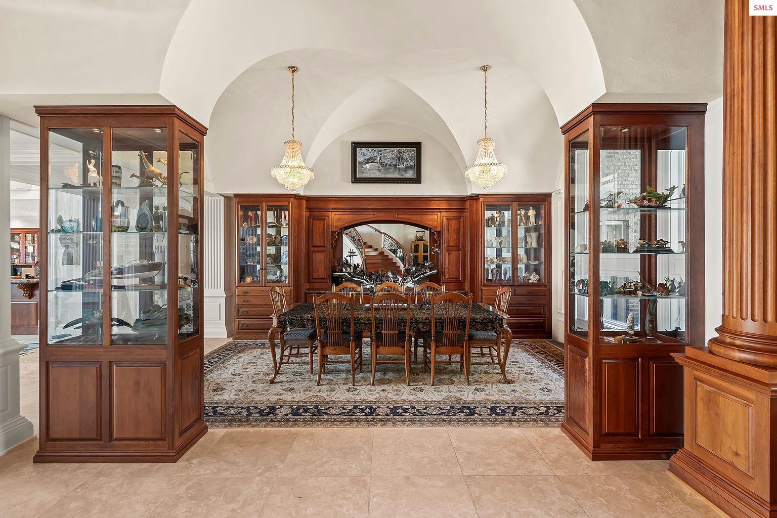 This is an elegant dining room featuring a long dining table with seating for ten, flanked by two large display cabinets filled with decorative items. The room has a high, arched ceiling with two chandeliers, and a large rug anchors the space. The overall impression is one of classic luxury and sophistication.