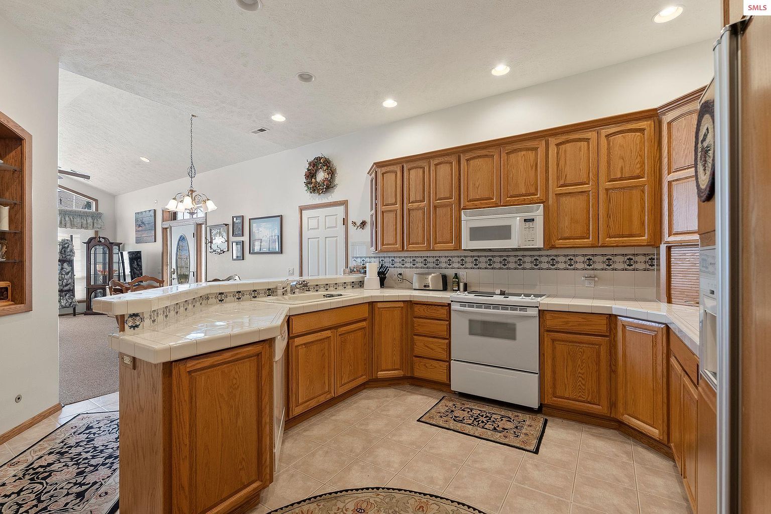 This is a well-lit kitchen featuring oak cabinetry, white appliances, and tiled countertops. The kitchen has a traditional style with a cozy rug in front of the oven and a decorative wreath on the wall. The perspective captures the kitchen's layout and connection to an adjacent living space.