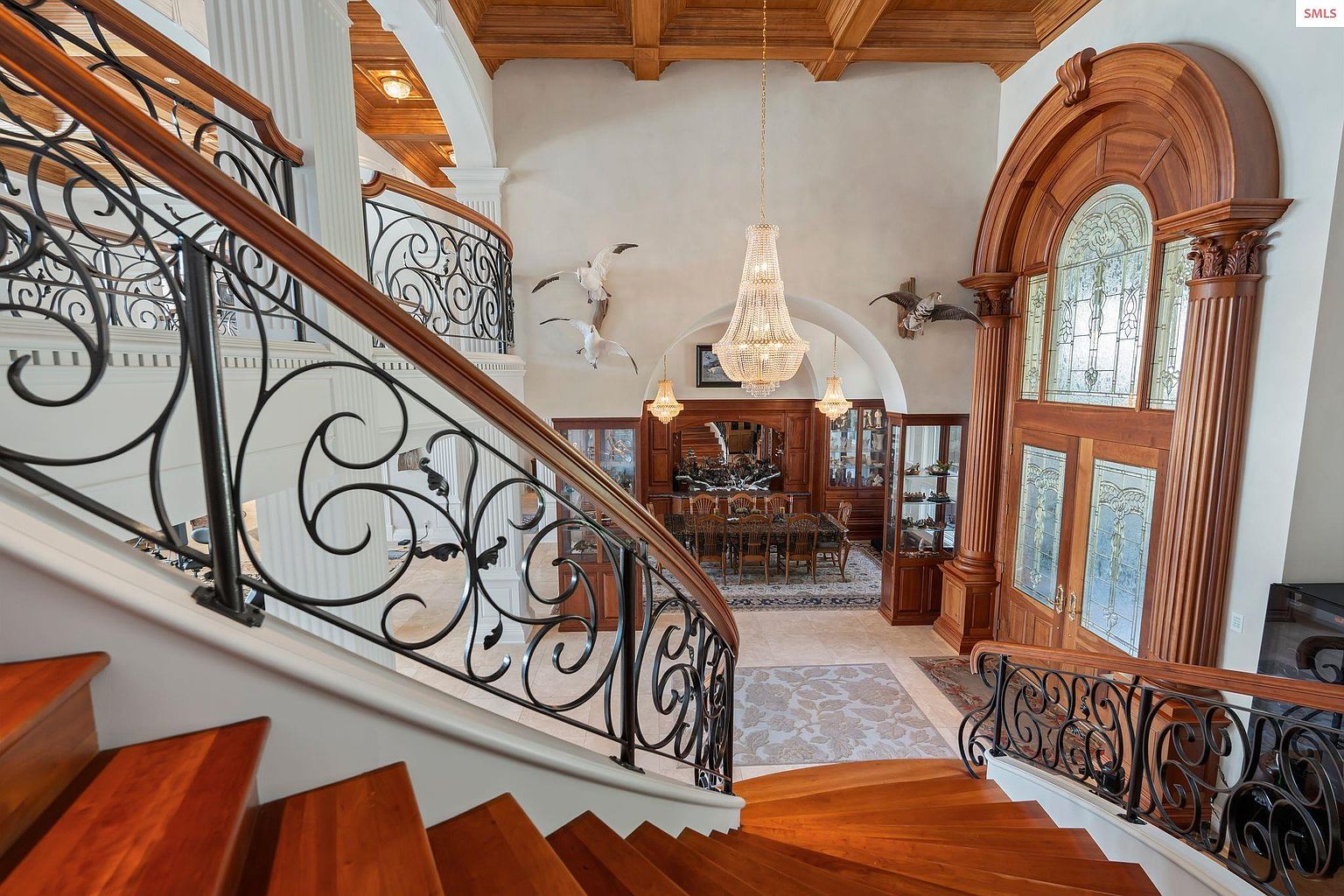 This grand foyer showcases a stunning staircase with ornate wrought iron railings and rich wood steps. A large, arched wooden doorway with decorative glass panels adds a touch of elegance, while a sparkling chandelier hangs above. The space is open and airy, creating a luxurious first impression.