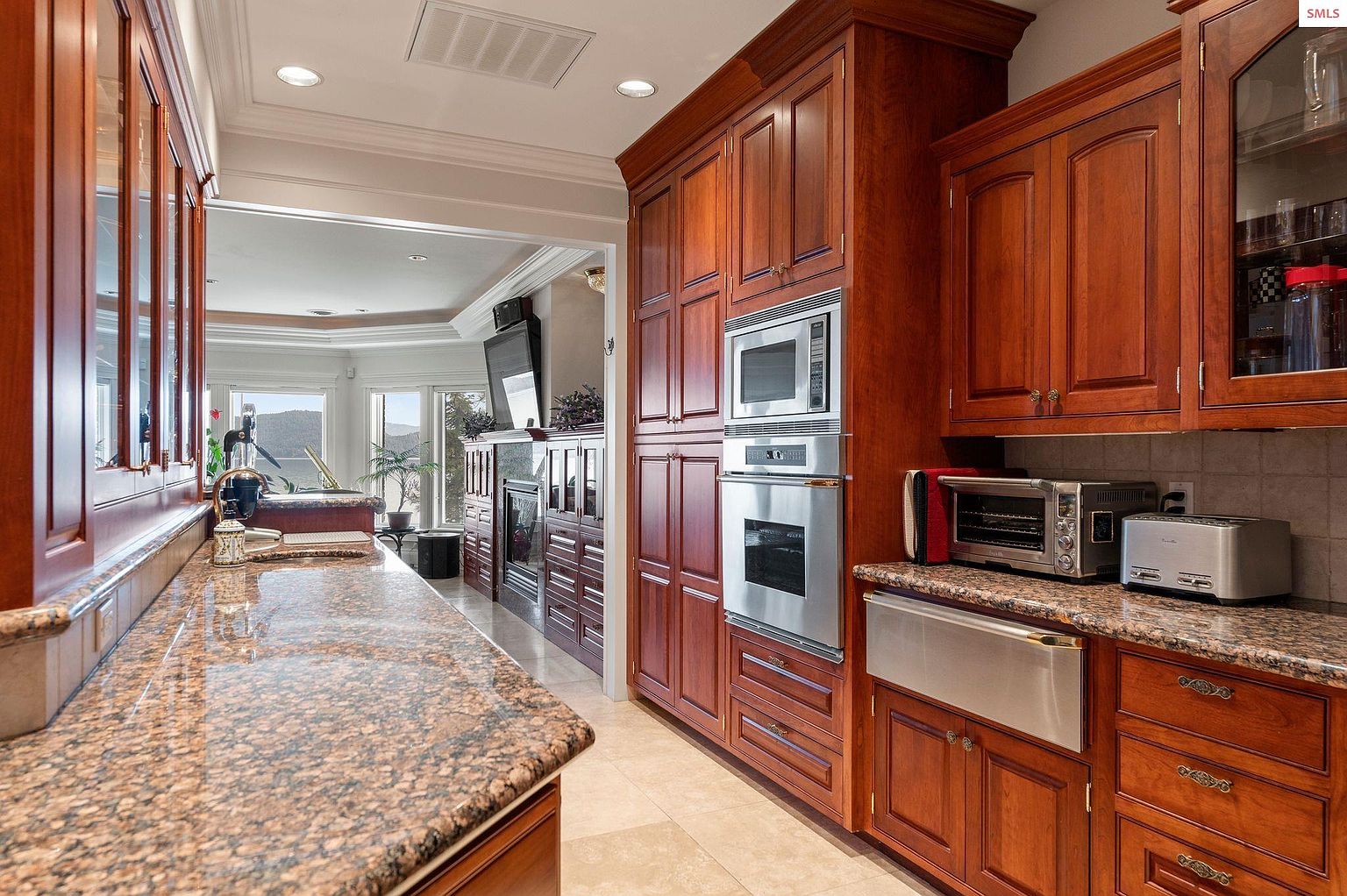 This is a well-appointed kitchen featuring rich, dark wood cabinetry, granite countertops, and stainless steel appliances. The kitchen has a built-in microwave and oven, along with a stainless steel sink and a toaster oven. The perspective is from the side, showcasing the depth and layout of the kitchen, with a glimpse into an adjacent living area.