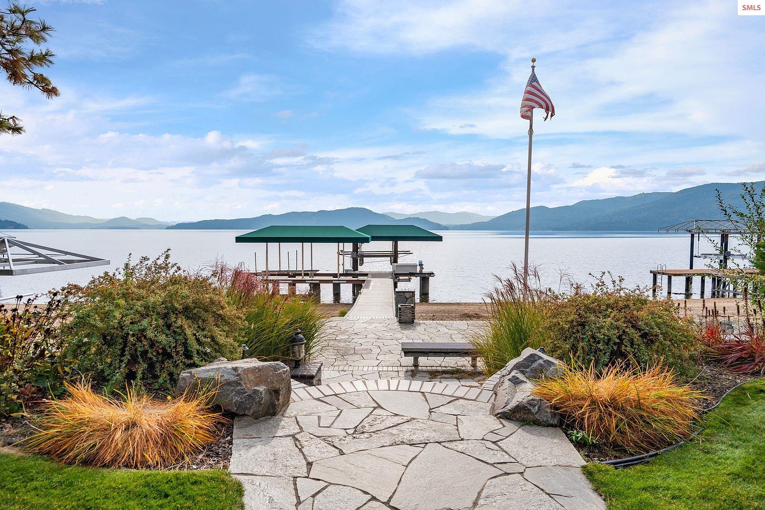This exterior shot showcases a beautifully landscaped yard leading to a private dock on a serene lake. A stone pathway, flanked by mature bushes and decorative rocks, guides the eye towards the dock featuring covered seating areas and an American flag. The scene evokes a sense of tranquility and upscale lakeside living, perfect for potential buyers seeking a peaceful retreat.