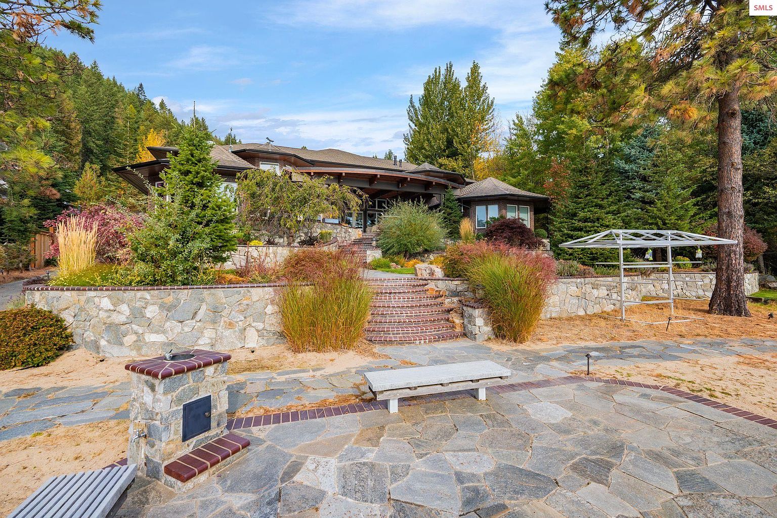 This image showcases a beautifully landscaped yard with a stone patio, brick accents, and lush greenery. A stone retaining wall adds dimension, while a pergola and outdoor fireplace enhance the outdoor living space. The overall impression is one of tranquility and sophisticated outdoor design, perfect for entertaining or relaxing.