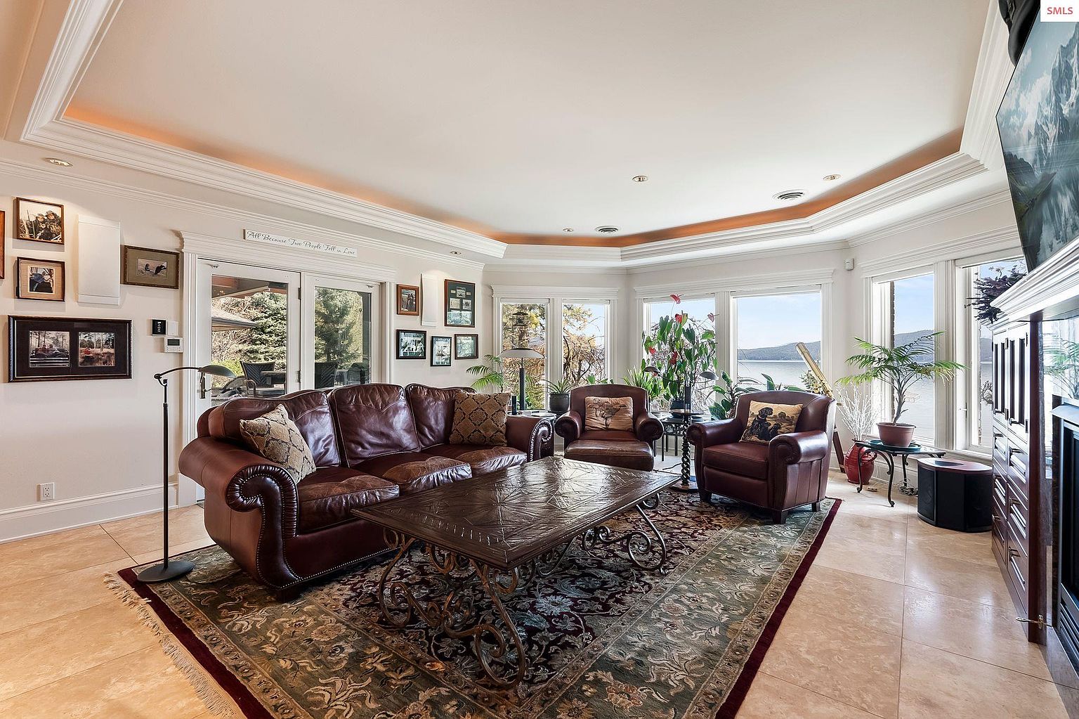 This is a luxurious living room featuring a large, plush leather sofa and two matching armchairs arranged around an ornate coffee table. The room is bathed in natural light from the surrounding windows, offering a view of the outdoors. An elegant rug anchors the seating arrangement, and various plants add a touch of nature to the space, creating a warm and inviting atmosphere.