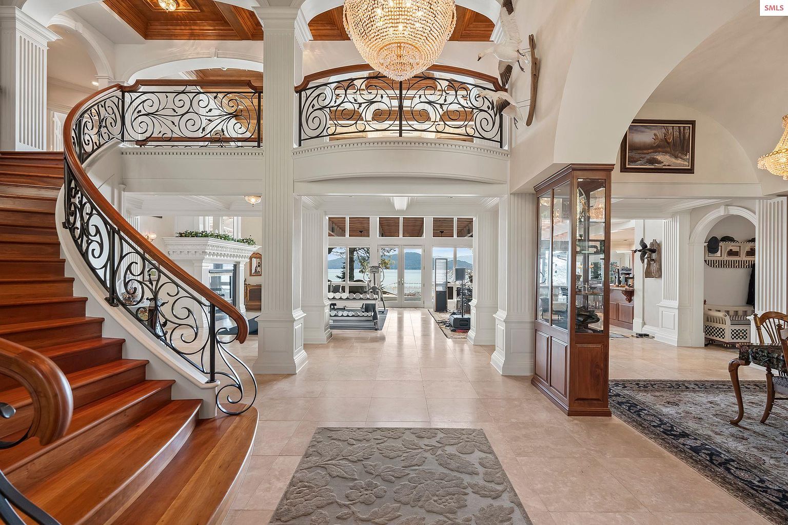 This grand foyer features a sweeping wooden staircase with ornate iron railings, leading to a second-floor balcony overlooking the entrance. A large crystal chandelier hangs from the coffered ceiling, illuminating the space. The flooring is light-colored tile, and the walls are adorned with architectural details such as columns and arched doorways, creating a luxurious and inviting atmosphere.