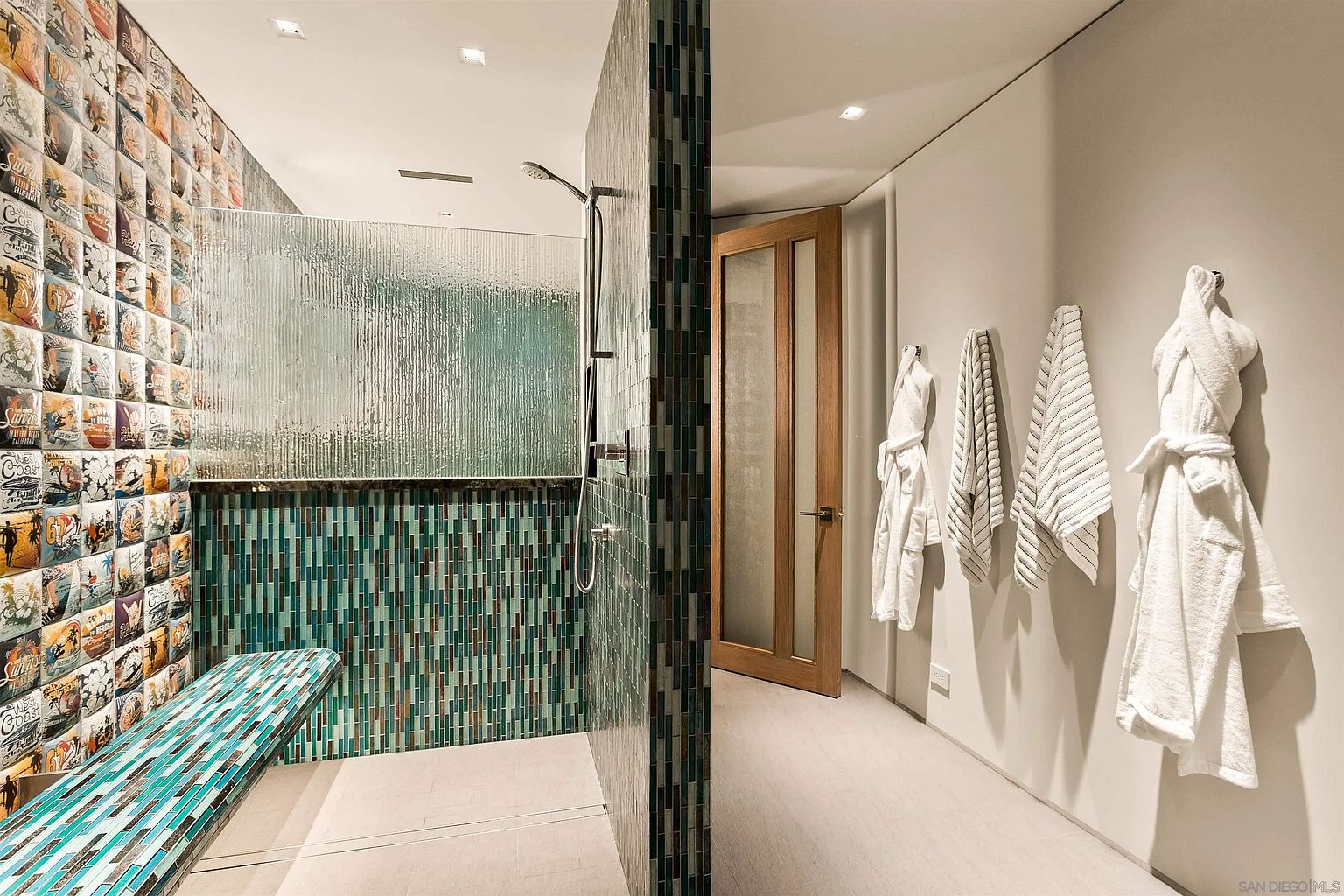 This modern bathroom features a striking walk-in shower with a vibrant, multi-colored vertical tile accent wall and a matching tiled bench. The space is complemented by a textured glass partition, a wooden door, and a clean, neutral wall adorned with several plush white robes hanging on hooks. The overall aesthetic is contemporary and spa-like, emphasizing unique textures and a bold color palette.