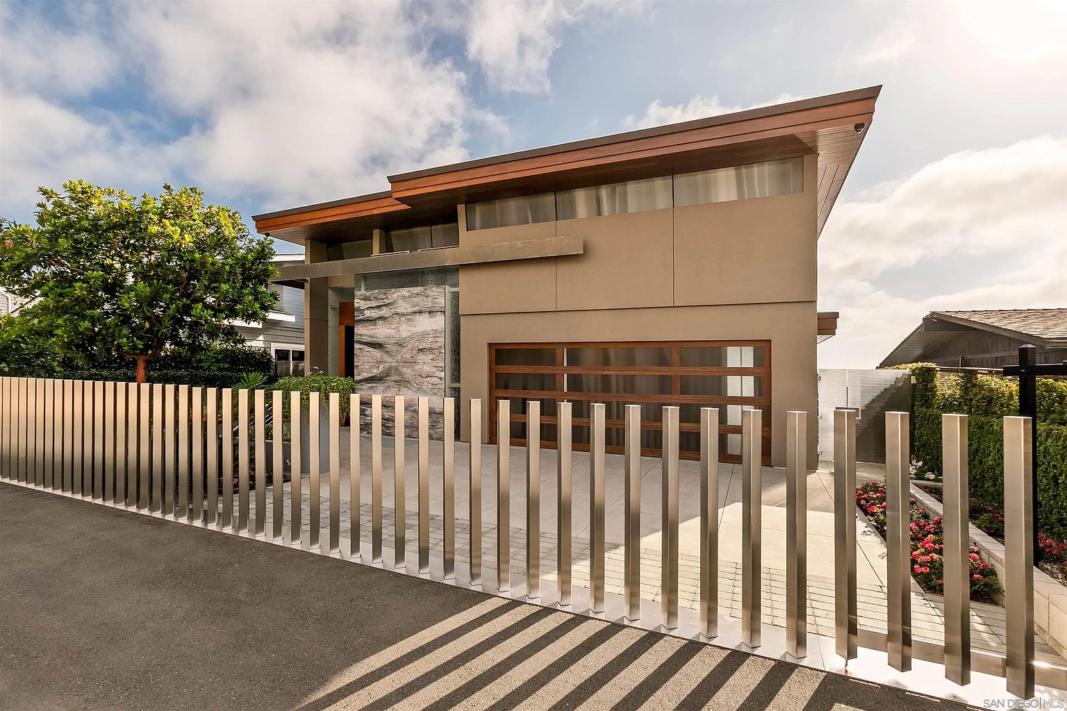 This modern residence features a striking facade with a prominent stone-clad accent wall and a multi-paneled garage door. A sleek, vertical metal slat fence provides a contemporary boundary, while the clean lines and warm wood-toned roofline emphasize the home's architectural sophistication. The perspective is a low-angle street view, capturing the interplay between the structured fencing and the home's geometric design.