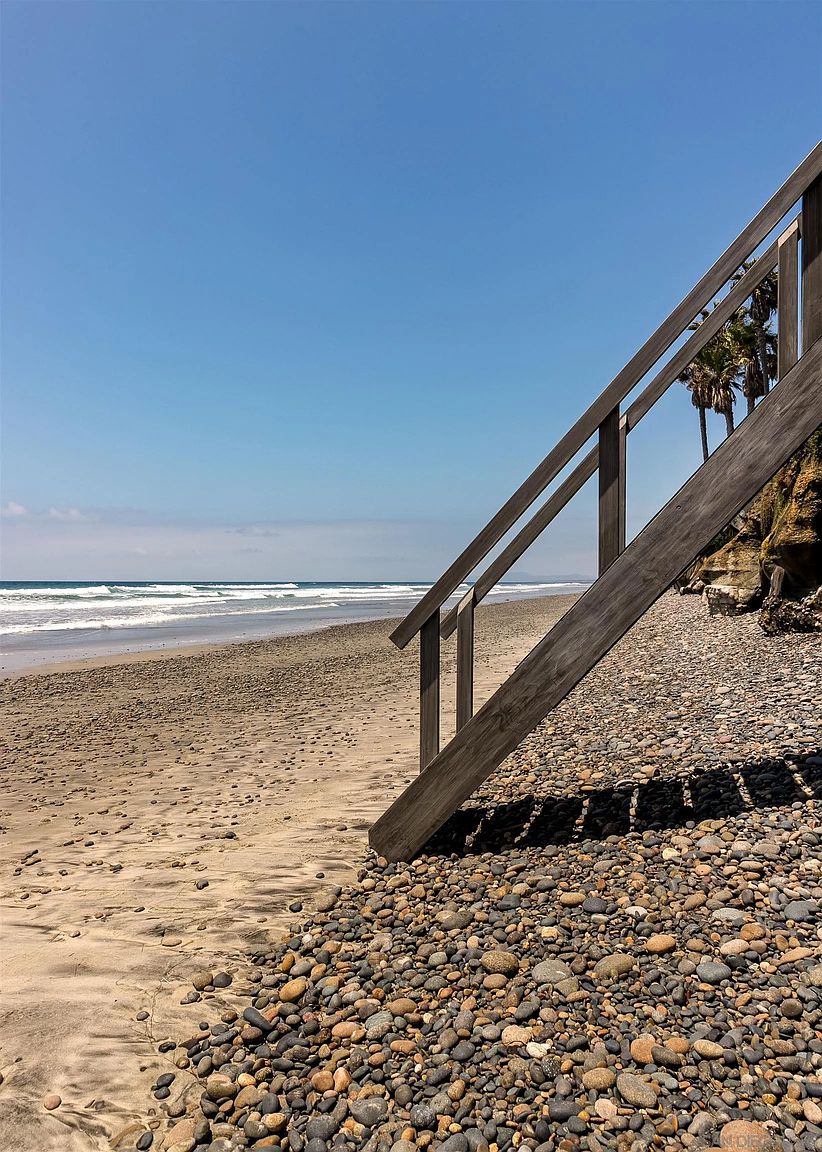 This image captures a wooden staircase leading directly down to a rocky beach, highlighting private or community access to the coastline. The perspective is from the top of the stairs looking out toward the vast ocean, emphasizing the natural beauty and proximity to the water. The scene features a clear blue sky, a sandy shoreline transitioning into pebbles, and coastal cliffs with palm trees in the background.
