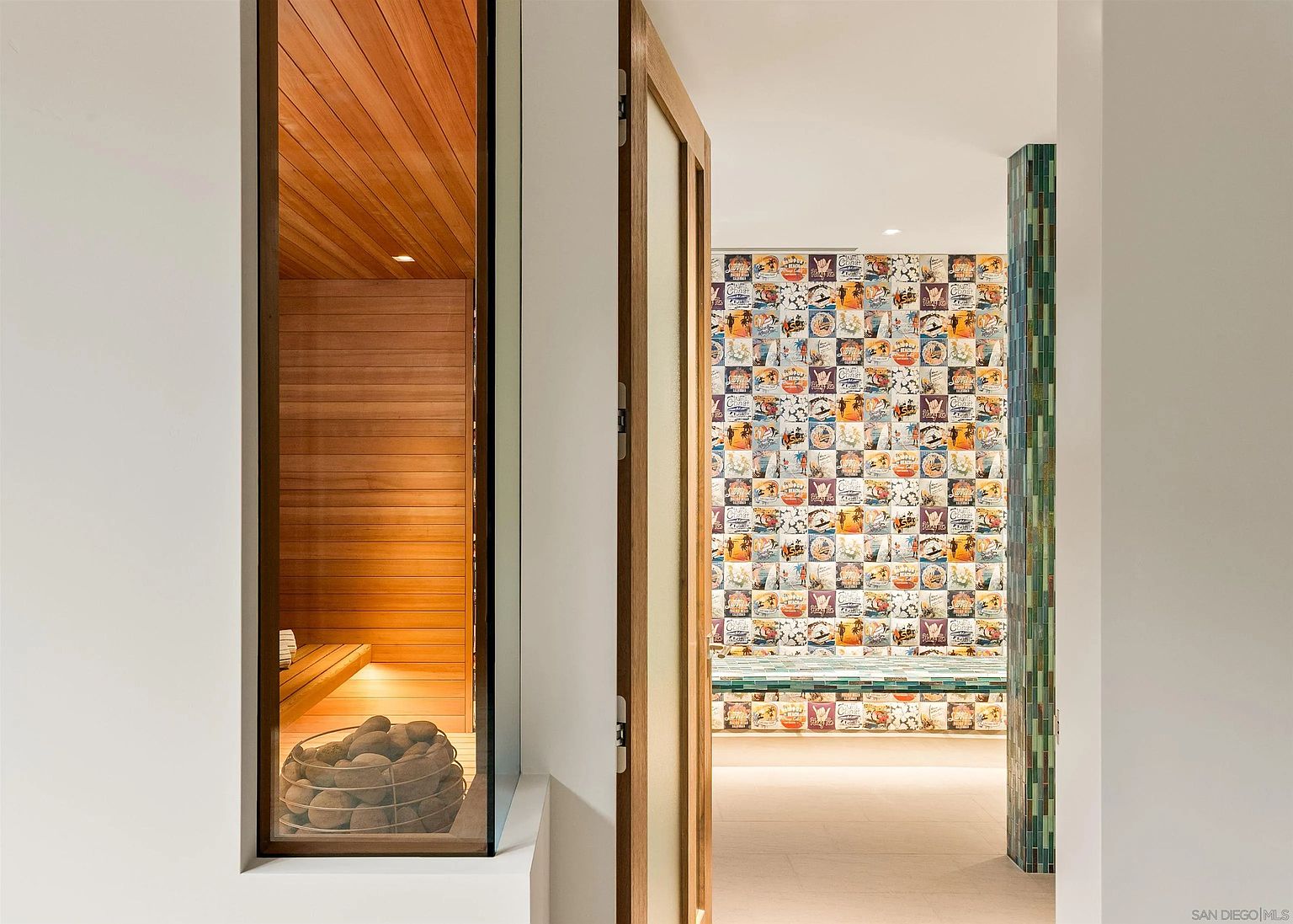 This image captures a unique transition space featuring a sauna on the left, visible through a large glass panel, and a vibrant, tiled wall on the right. The sauna is finished in warm, light wood with a basket of stones in the foreground, while the adjacent area showcases a wall covered in colorful, vintage-style graphic tiles. The perspective looks through a doorway, highlighting the contrast between the serene, natural wood tones of the sauna and the eclectic, artistic flair of the tiled feature wall.
