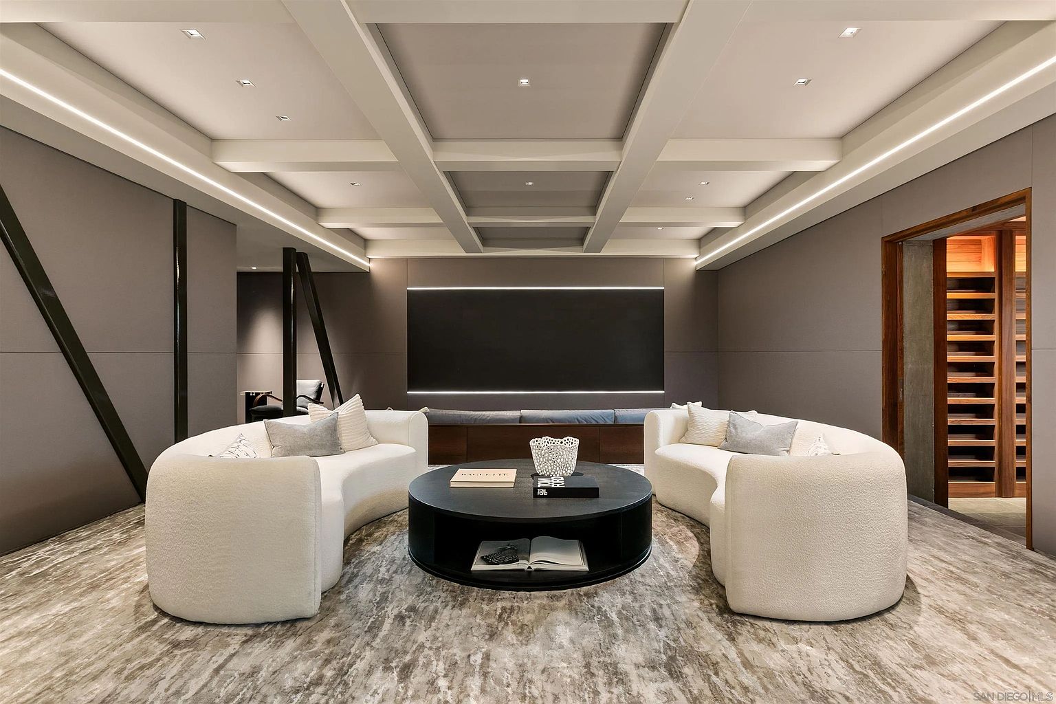 This sophisticated media room features a symmetrical layout with two curved, cream-colored sofas facing a central circular coffee table. A large, dark-toned screen is mounted on the wall, accented by integrated lighting, while the ceiling showcases a detailed coffered design. To the right, a glimpse of a wine storage area adds a touch of luxury to this modern, neutrally-toned entertainment space.