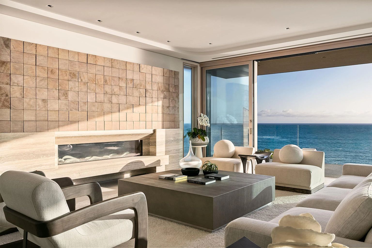 This sophisticated living room features a textured wood-block accent wall and a sleek, linear fireplace set within a stone surround. The space is furnished with plush, neutral-toned seating and a large, low-profile coffee table, all oriented toward expansive glass sliding doors that reveal a stunning, unobstructed ocean view. The room exudes a serene, high-end coastal aesthetic, emphasizing natural light and seamless indoor-outdoor integration.