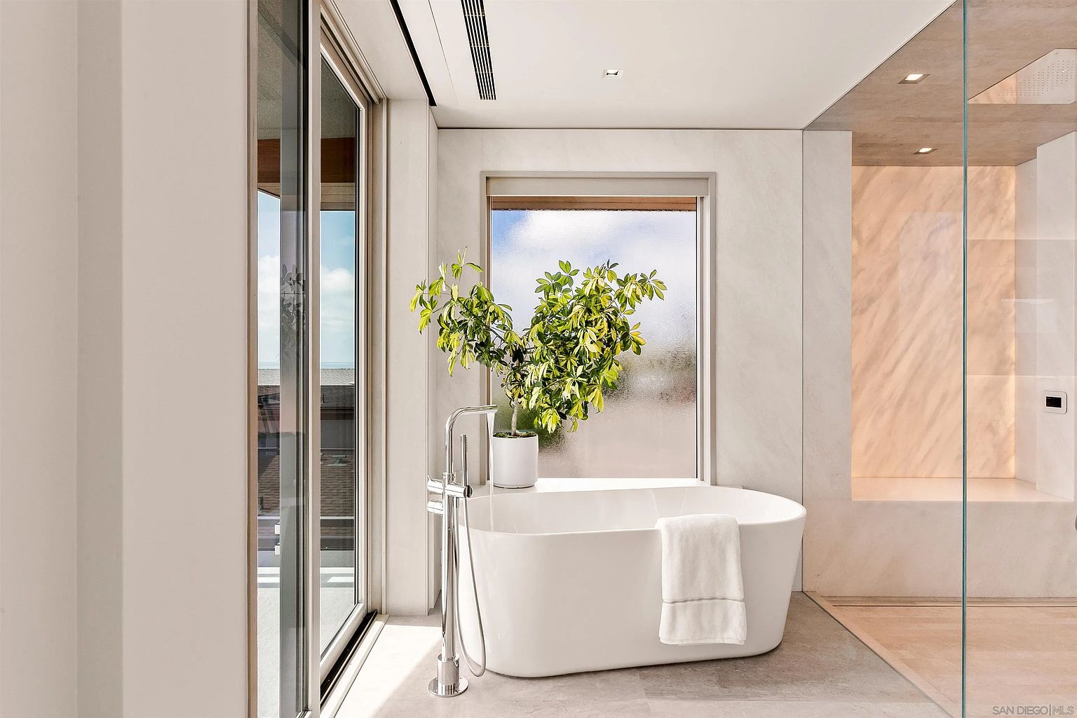 This luxurious primary bathroom features a sleek, freestanding soaking tub positioned beneath a large window that brings in natural light. To the right, a glass-enclosed walk-in shower with light-toned stone walls creates a seamless, spa-like aesthetic. The room is accented by a potted plant on the tub ledge and large-format floor tiles, offering a bright and modern atmosphere.