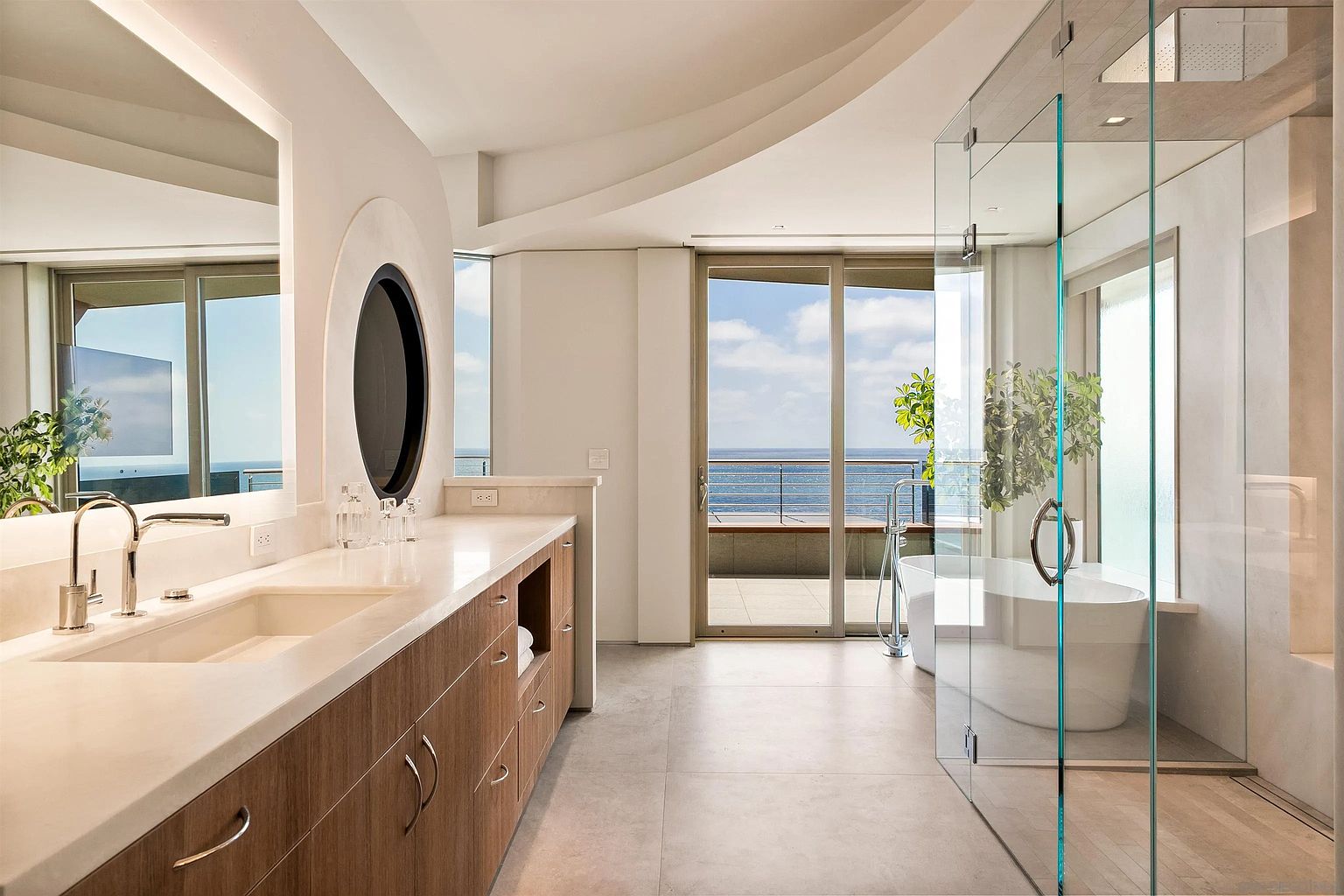 This luxurious primary bathroom features a sleek vanity with a light-toned countertop and wood-finish cabinetry, complemented by a unique oval mirror. A glass-enclosed shower area sits adjacent to a freestanding soaking tub, which is positioned to take advantage of the expansive ocean view through the large sliding glass doors. The space is finished with neutral, large-format floor tiles, creating a serene and modern atmosphere.