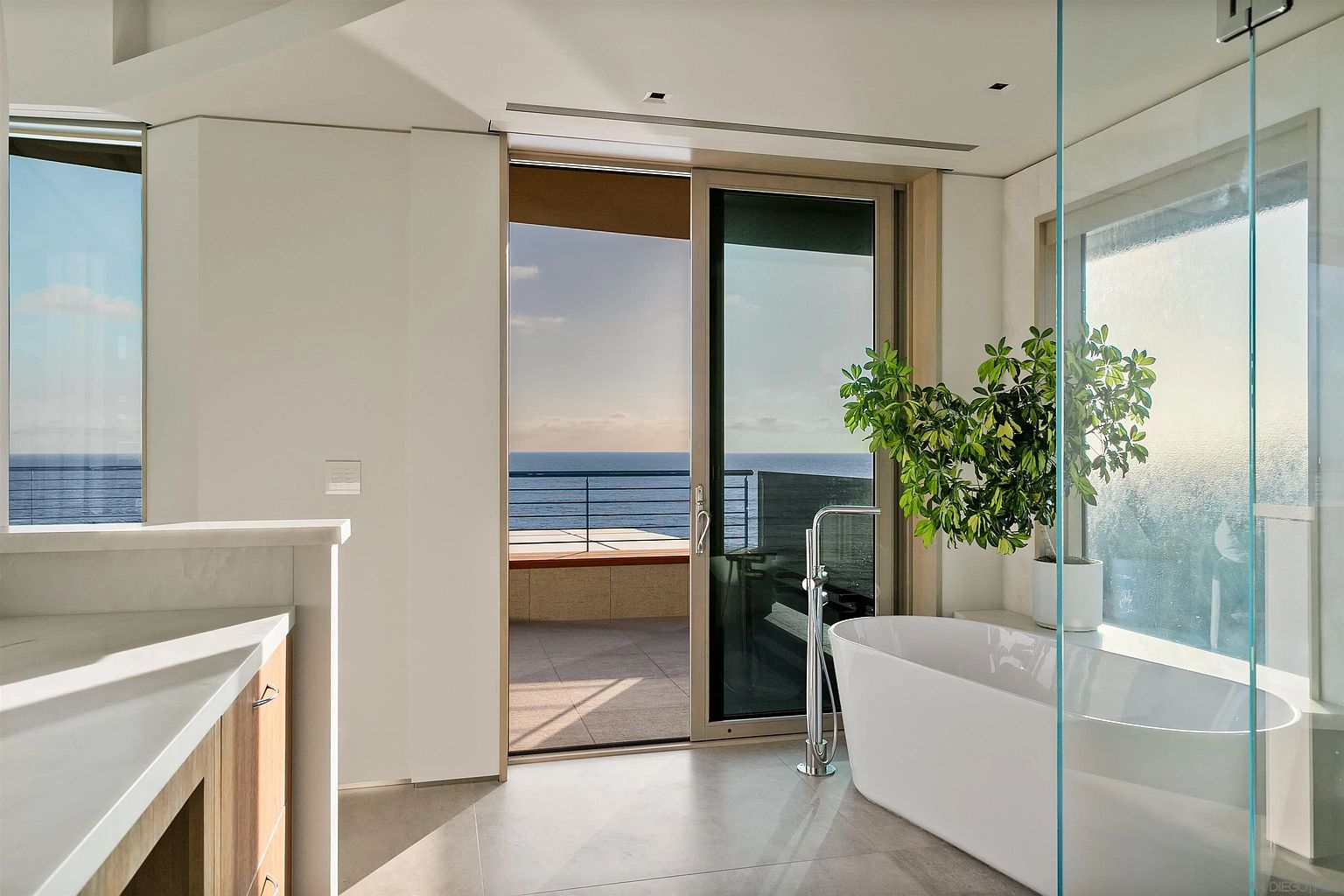 This modern primary bathroom features a sleek, freestanding soaking tub positioned next to a large glass-enclosed shower and a sliding glass door that opens to a private balcony. The space is bathed in natural light, highlighting the neutral stone flooring and the expansive ocean view beyond the balcony railing. A potted plant adds a touch of greenery, enhancing the serene, spa-like atmosphere of this coastal retreat.