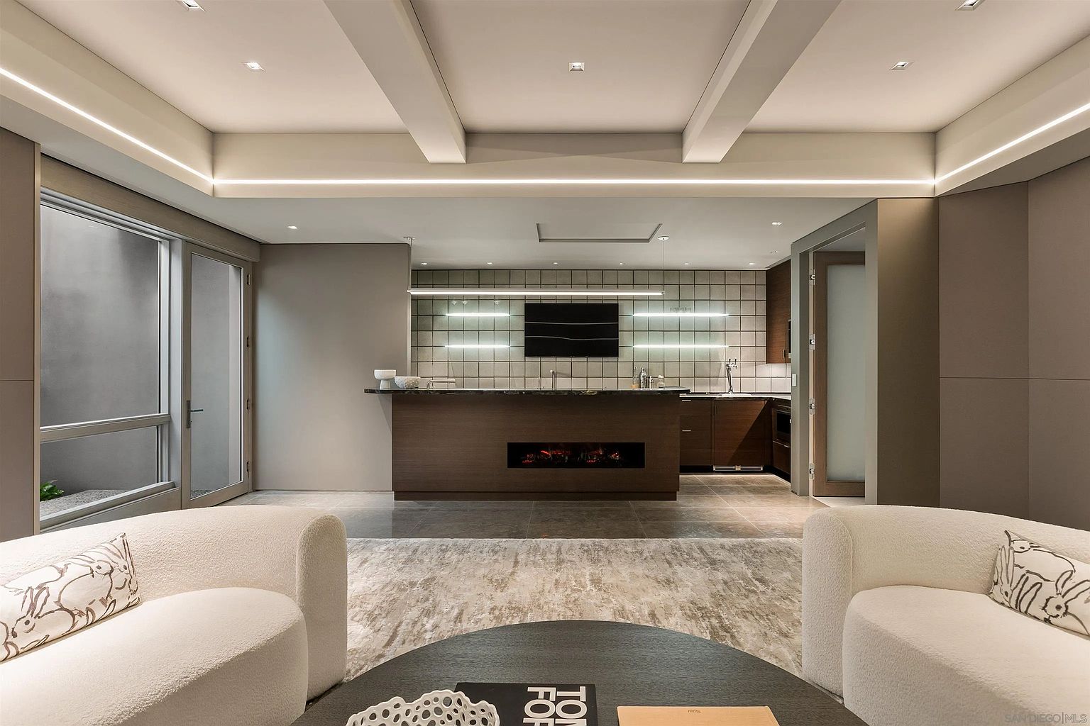 This sophisticated living area features a central bar with a built-in fireplace, serving as a focal point between the seating area and the kitchen. The room is defined by a neutral color palette, recessed ceiling lighting, and large windows that provide a view to the exterior. The space is furnished with two curved, light-colored armchairs and a dark circular coffee table, creating an inviting and modern atmosphere.