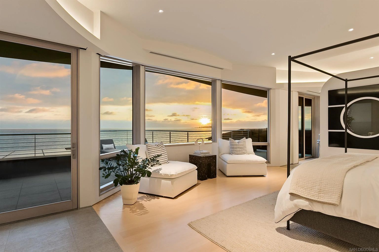 This luxurious primary bedroom features a canopy bed and two plush white lounge chairs positioned to take advantage of the expansive ocean views through large windows. The room is bathed in the warm, golden light of a sunset, creating a serene and sophisticated atmosphere. A neutral color palette and minimalist decor emphasize the seamless connection between the interior space and the outdoor balcony.