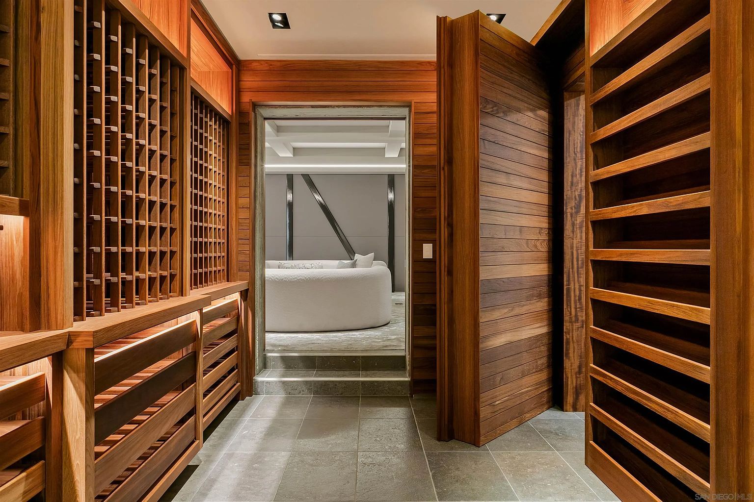 This sophisticated wine cellar features extensive custom wood shelving designed for bottle storage, creating a warm and textured aesthetic. A large, heavy wooden door stands partially open, revealing a transition into an adjacent living space with a curved sofa. The room is finished with neutral stone-look floor tiles and recessed lighting, offering a refined and climate-controlled atmosphere.