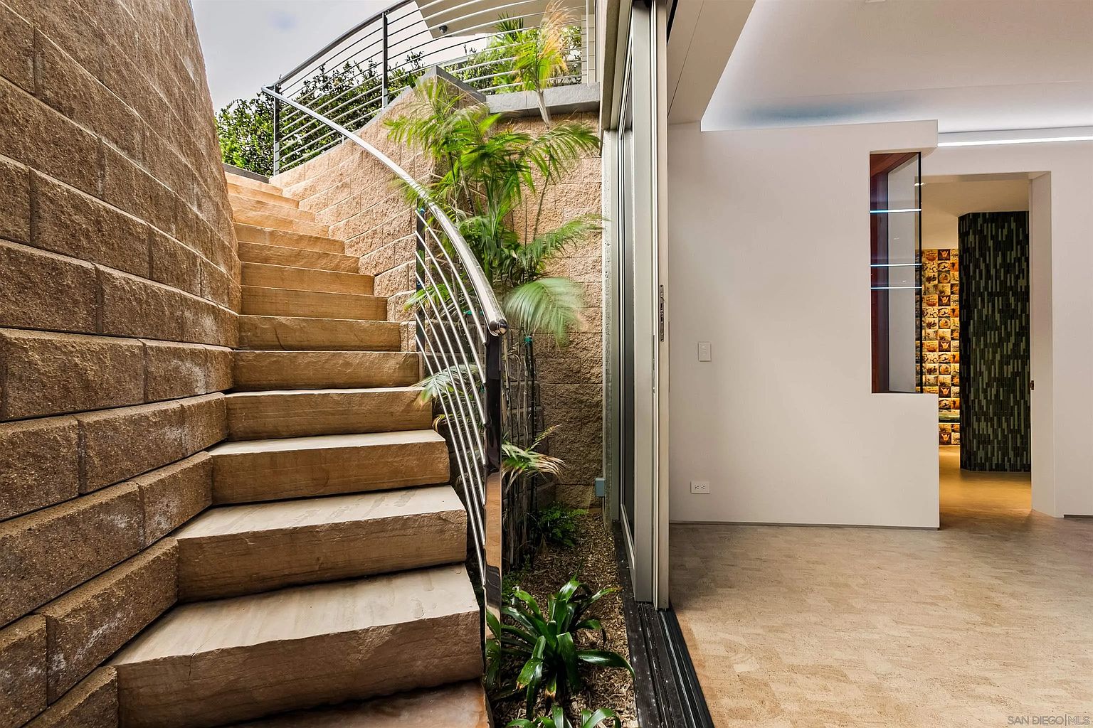 This unique transition space features a curved stone staircase leading upward, flanked by a textured stone retaining wall and a sleek metal railing. To the right, a large glass sliding door opens into a bright interior area with light-toned flooring and a decorative wall feature. The integration of an indoor-outdoor garden space at the base of the stairs creates a sophisticated, architectural aesthetic.