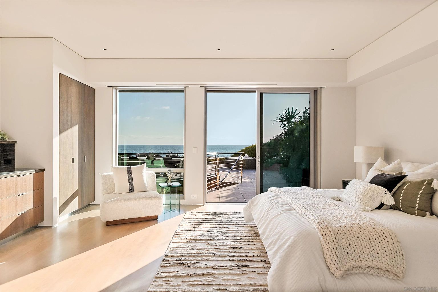 This bright and airy primary bedroom features a minimalist aesthetic with light wood flooring and neutral walls. A large bed with layered white linens and textured pillows serves as the focal point, while a plush white armchair sits near the expansive windows that offer a direct view of the ocean. The room opens onto a private patio, seamlessly blending indoor and outdoor living spaces with a clean, contemporary design.