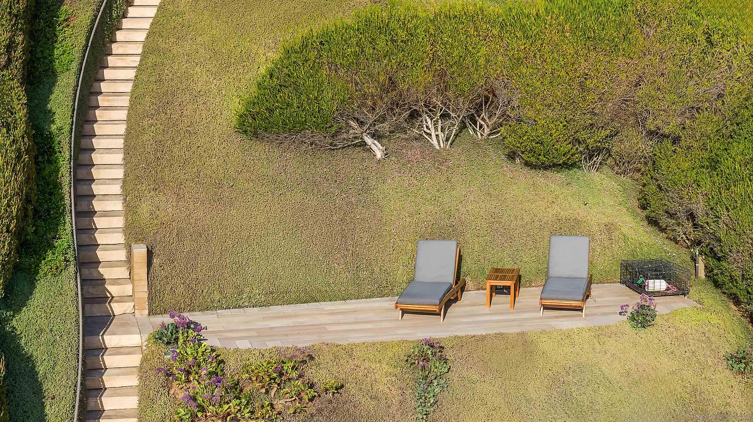 This aerial perspective captures a landscaped hillside garden featuring a stone-paved patio area furnished with two lounge chairs and a small side table. A winding stone staircase descends along the side of the grassy slope, framed by lush greenery and flowering plants. The scene offers a serene and private outdoor retreat, emphasizing the property's well-maintained terraced grounds.
