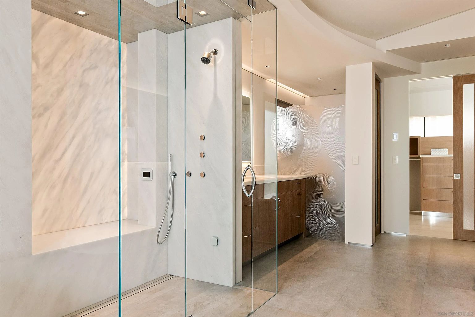 This modern primary bathroom features a spacious walk-in shower enclosed by floor-to-ceiling glass panels, showcasing a built-in stone bench and wall-mounted shower fixtures. Adjacent to the shower is a vanity with wood-toned cabinetry and a decorative, textured glass privacy screen that adds a sculptural element to the room. The perspective captures the clean lines and high-end finishes of the space, leading the eye toward an open doorway that reveals a walk-in closet area.