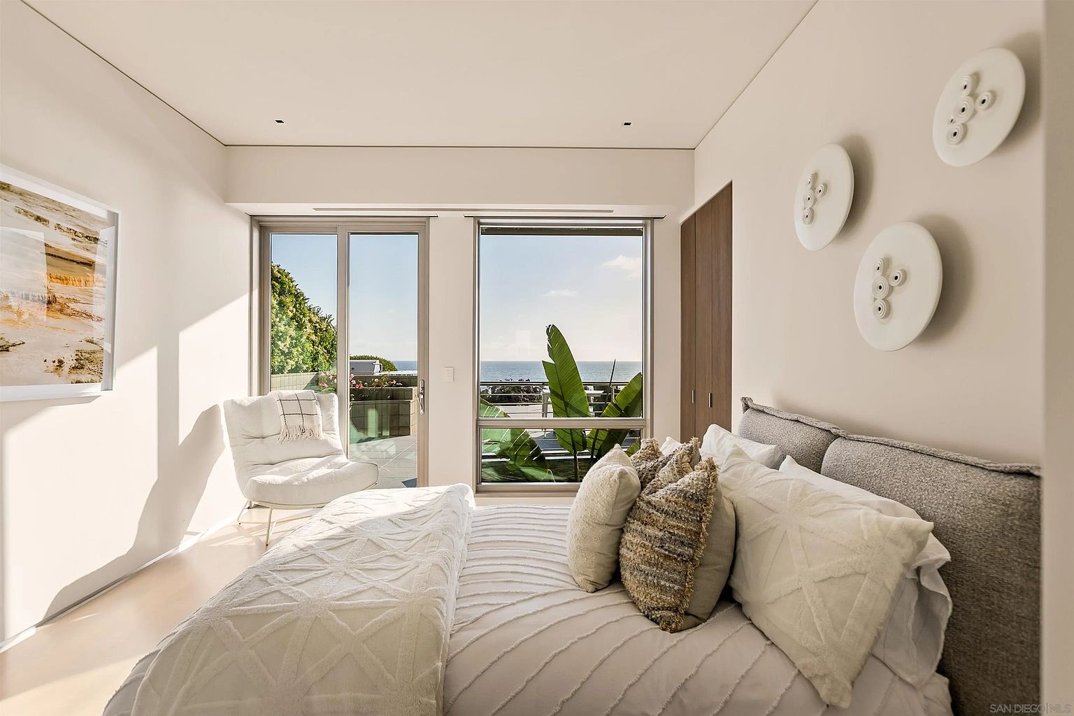 This serene primary bedroom features a plush bed with textured bedding and a neutral color palette, creating a calm and inviting atmosphere. Large windows provide an expansive view of the ocean and a private balcony, while unique circular wall art adds a modern decorative touch. The room is bathed in natural light, highlighting the clean lines and minimalist aesthetic of the space.