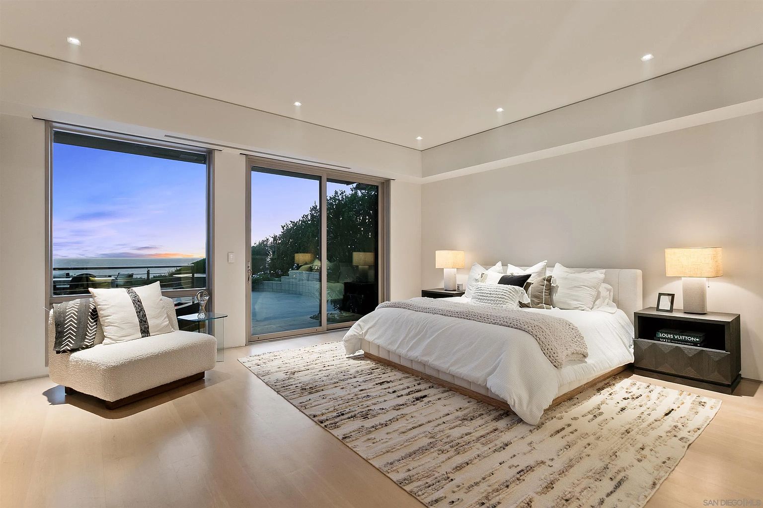 This spacious primary bedroom features a neutral color palette and a contemporary design, centered around a large bed with plush white bedding. Large sliding glass doors provide an expansive view of the ocean and outdoor patio, filling the room with natural light. The space is accented by a textured area rug, minimalist bedside tables with warm lamps, and a comfortable accent chair, creating a serene and luxurious atmosphere.