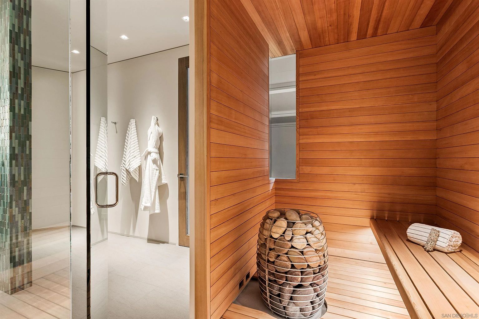 This image showcases a luxurious sauna room featuring warm-toned wood paneling on the walls and ceiling, complemented by a slatted wood bench. A cylindrical metal heater filled with smooth stones serves as the focal point, while an adjacent glass-enclosed shower area reveals white robes hanging on the wall. The clean, minimalist design creates a serene and spa-like atmosphere perfect for relaxation.