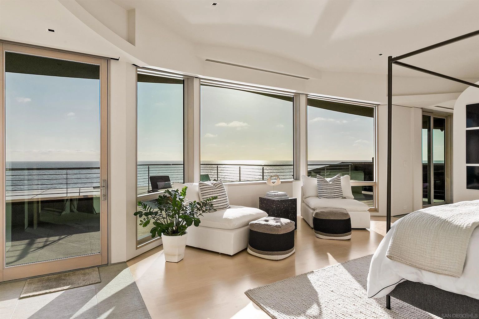 This primary bedroom features a sophisticated, modern aesthetic with light wood flooring and a neutral color palette. Large windows provide expansive, unobstructed views of the ocean, while a pair of white lounge chairs and matching ottomans create a serene seating area. A four-poster bed frame is partially visible on the right, and a potted plant adds a touch of greenery to the bright, airy space.