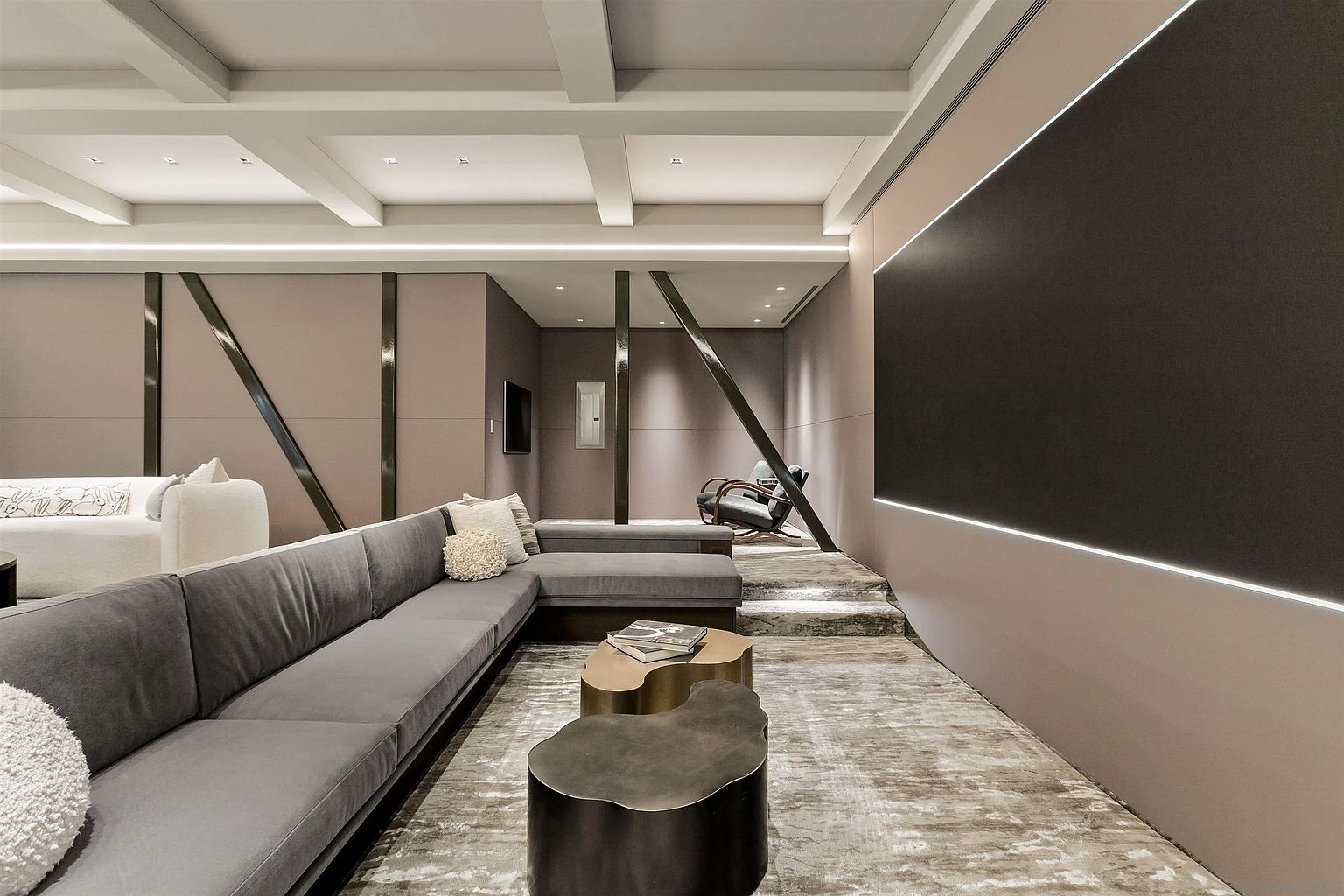 This sophisticated media room features a large, plush grey sectional sofa arranged around unique, organic-shaped coffee tables. The space is defined by a coffered ceiling, a raised platform area with additional seating, and a prominent, dark-toned wall accented with integrated linear lighting. The room's modern aesthetic is enhanced by structural diagonal beams and a neutral, textured color palette that creates a cozy, cinematic atmosphere.
