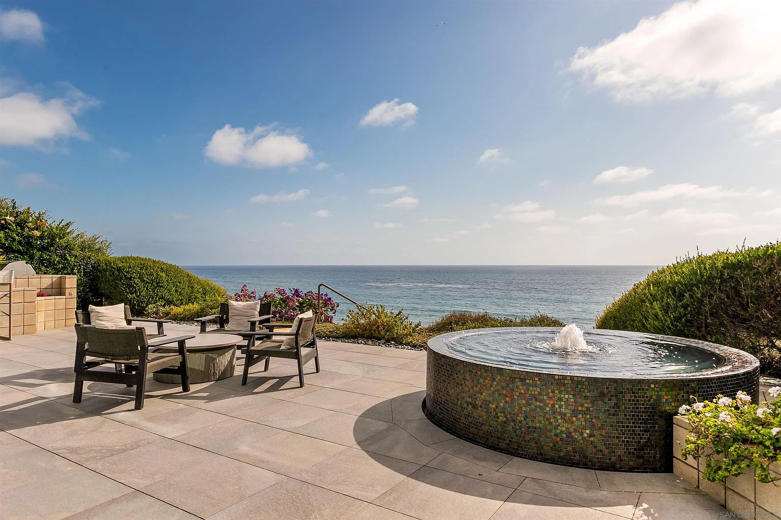 This expansive oceanfront patio features a circular tiled fountain as a focal point, set against a backdrop of the Pacific Ocean. The space is furnished with a modern outdoor seating arrangement, including a circular fire pit table surrounded by comfortable chairs. The scene is captured from a slightly elevated perspective, emphasizing the seamless transition between the paved terrace and the coastal horizon.