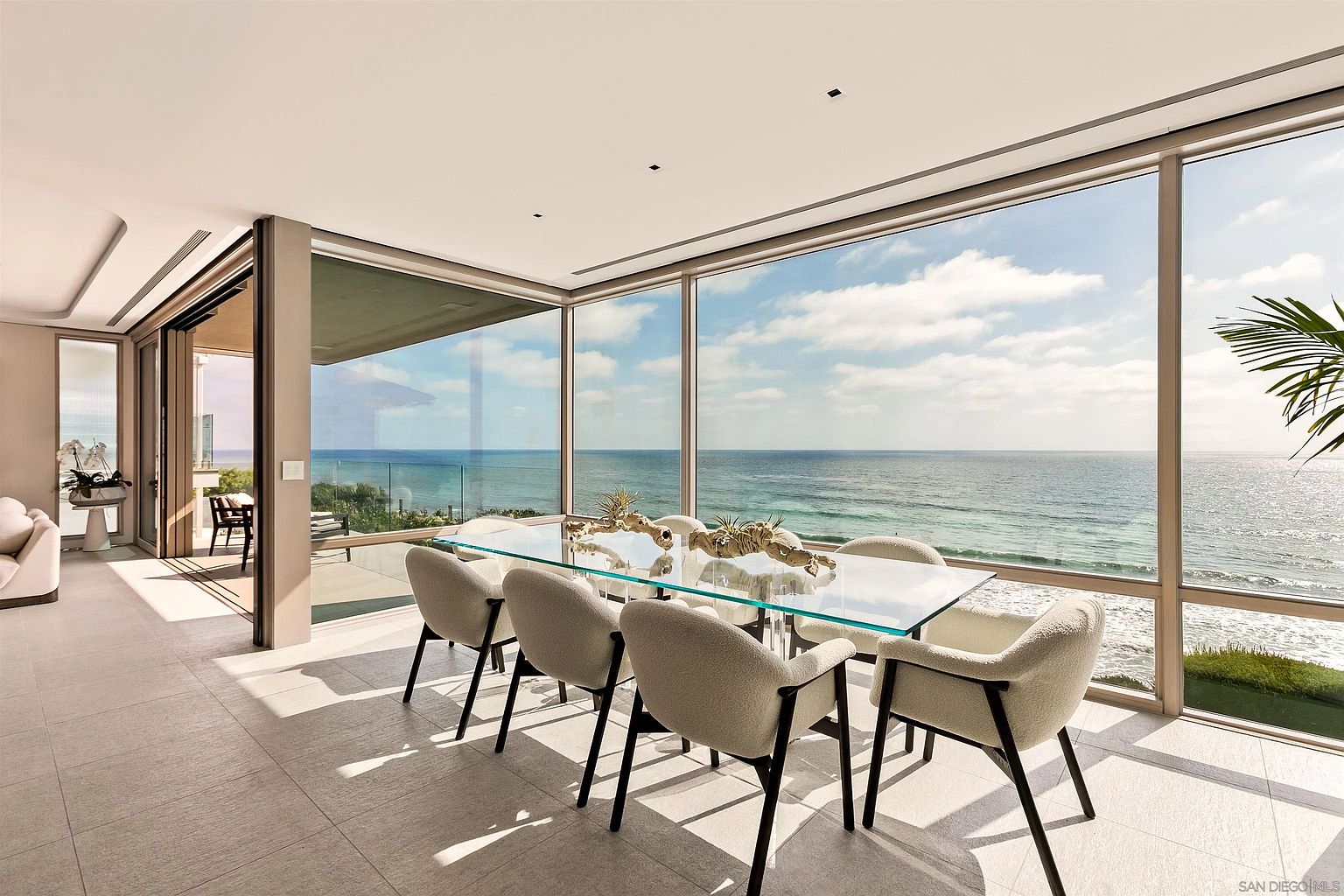 This bright and airy dining room features a glass-topped table surrounded by six modern, light-colored upholstered chairs. The space is defined by expansive floor-to-ceiling windows that provide an unobstructed, panoramic view of the ocean and coastline. The minimalist design, neutral color palette, and light-toned tile flooring create a serene and sophisticated atmosphere.