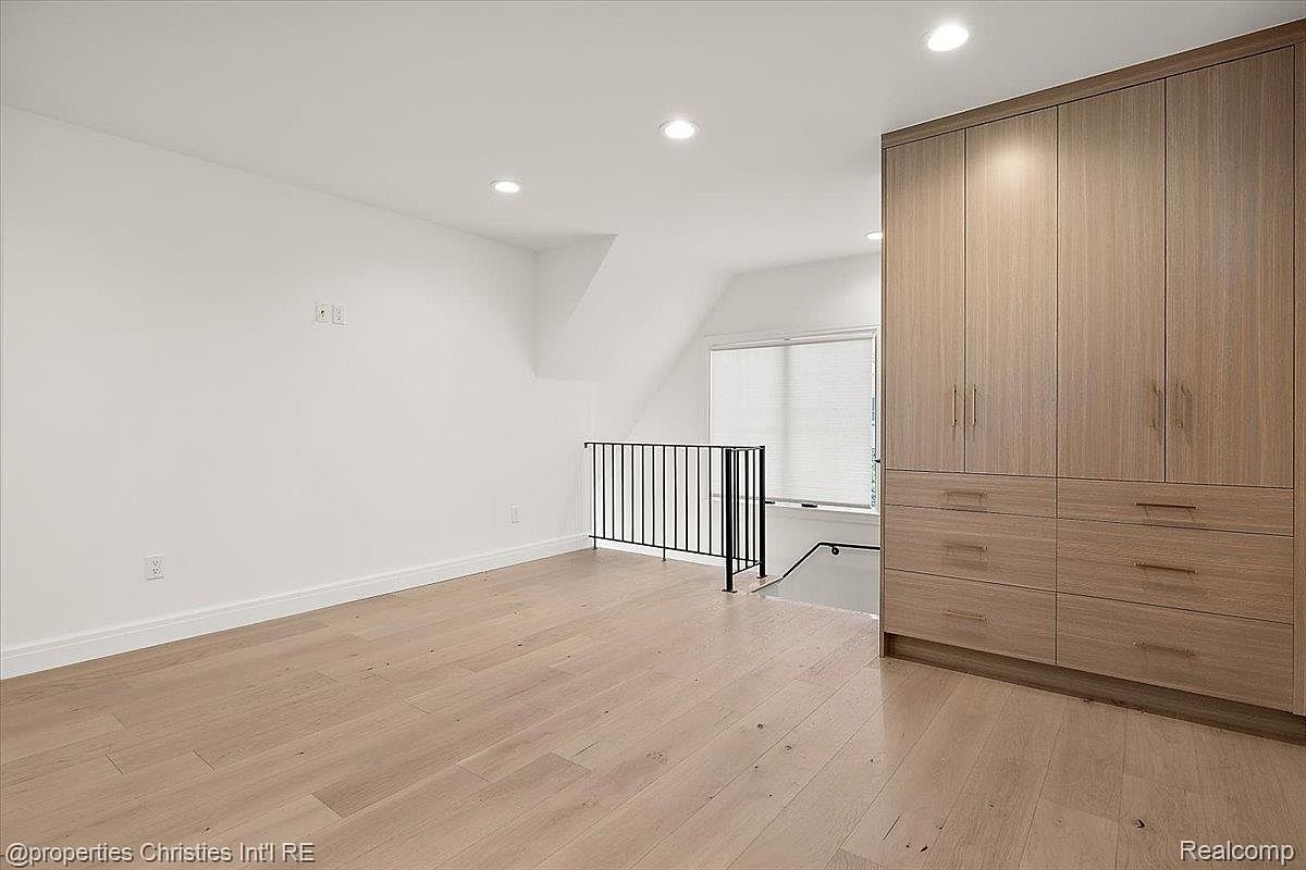 This is an interior shot of a bedroom featuring light hardwood floors and white walls. A large, built-in wooden wardrobe stands against one wall, offering ample storage. A black railing indicates a staircase or landing, adding architectural interest to the space.