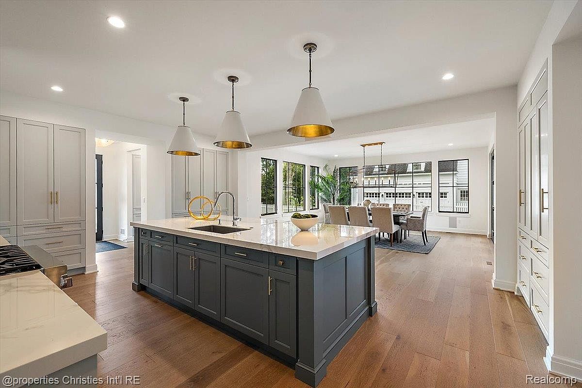This is a bright and spacious kitchen featuring a large center island with a dark gray base and a light countertop. The kitchen includes modern pendant lighting, light gray cabinetry, and hardwood flooring. The open layout connects to a dining area with large windows, creating an inviting and functional space.