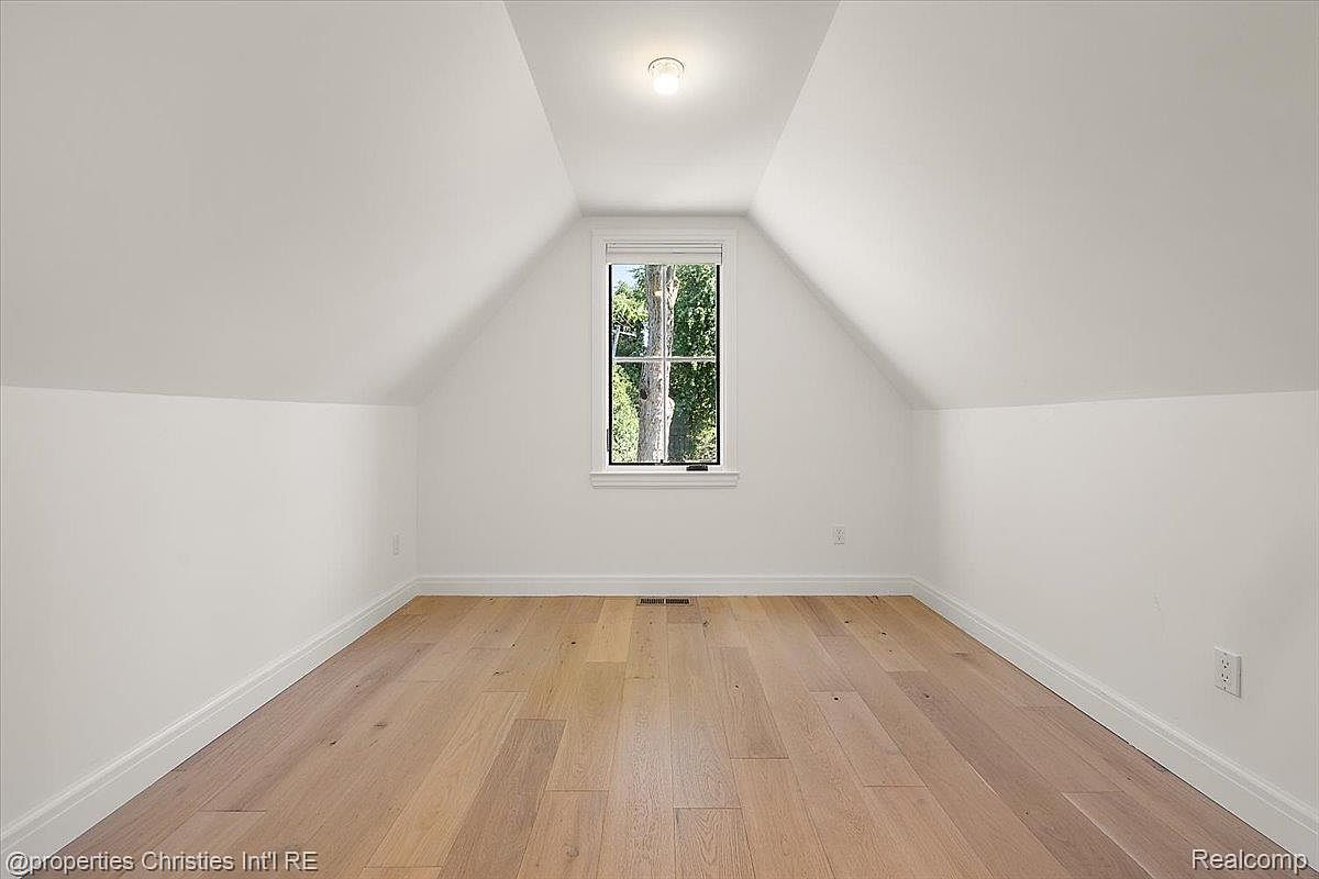 The image showcases a bright, empty bedroom with an attic-style ceiling and light hardwood flooring. A single window provides natural light and a view of trees outside. The room is painted in a neutral white, creating a clean and spacious feel, ideal for a guest bedroom or flexible living space.