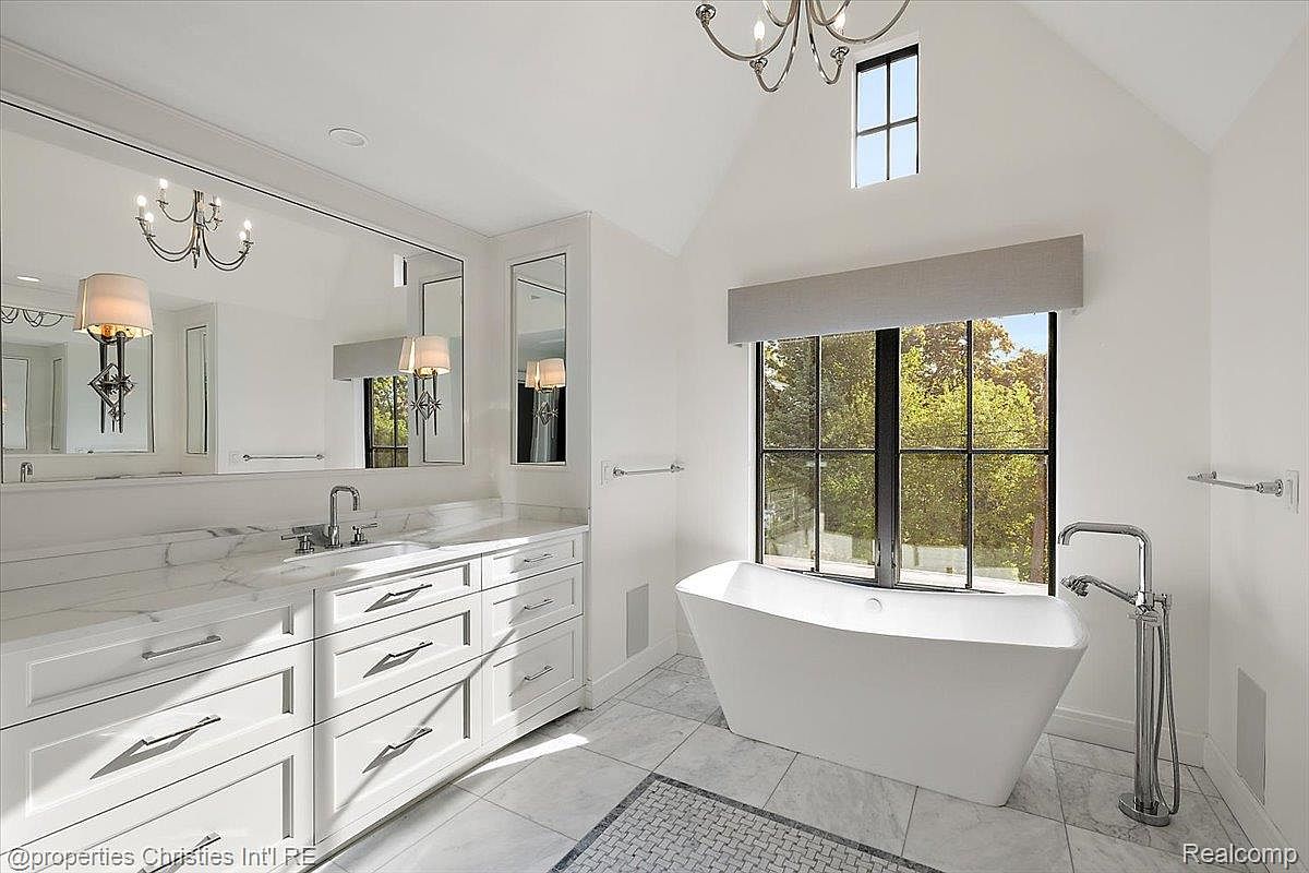 This is a luxurious primary bathroom featuring a large white freestanding bathtub positioned in front of a window with a view of trees. The bathroom also includes a spacious vanity with white cabinetry, marble countertops, and a large mirror. The overall style is elegant and bright, creating a spa-like atmosphere.