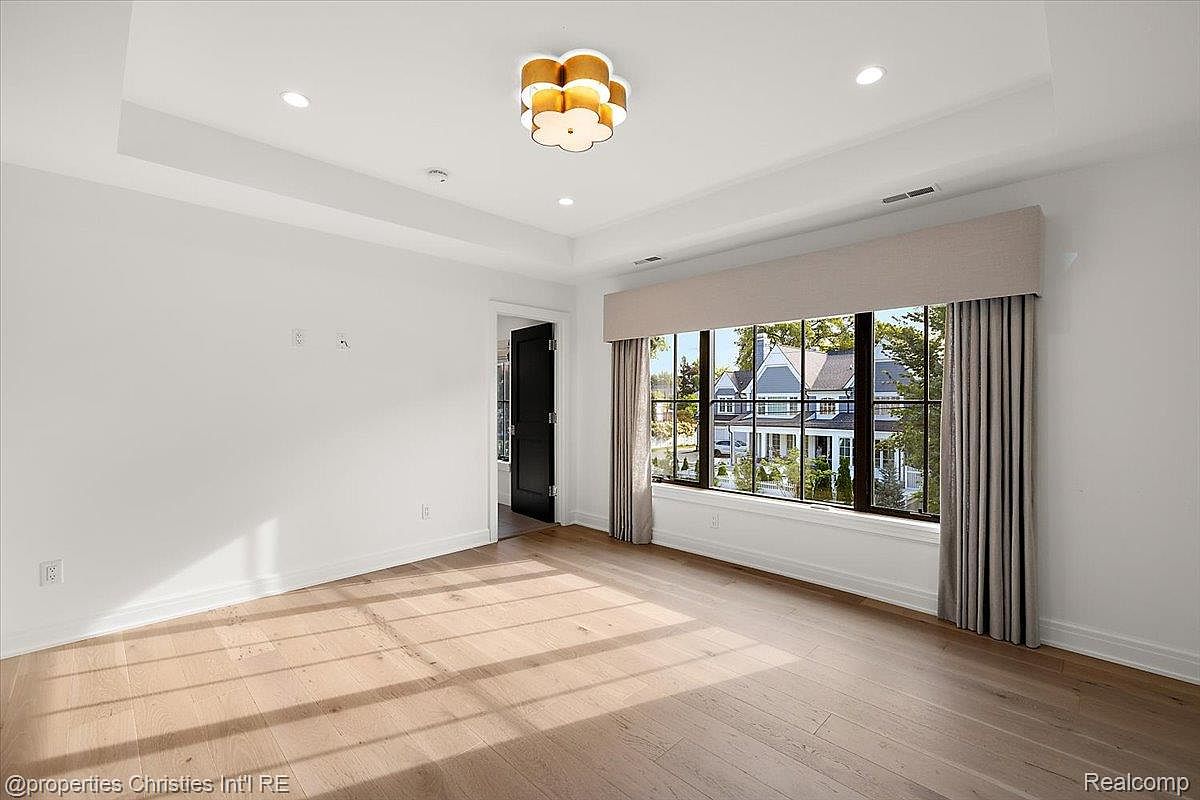 This is a bright and spacious primary bedroom featuring hardwood floors and a large window with neutral-toned curtains. The room has white walls and a modern, decorative light fixture. The overall impression is clean, airy, and inviting, suggesting a comfortable and luxurious living space.