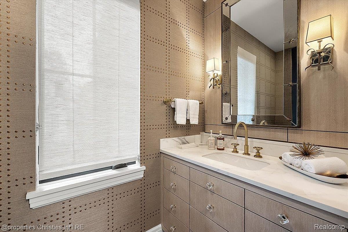 This is a well-appointed guest bathroom featuring a stylish vanity with a marble countertop and crystal knobs. The walls are covered in a textured wallpaper, and a framed mirror hangs above the sink, reflecting the light from the sconces. Rolled towels and decorative items add a touch of luxury.