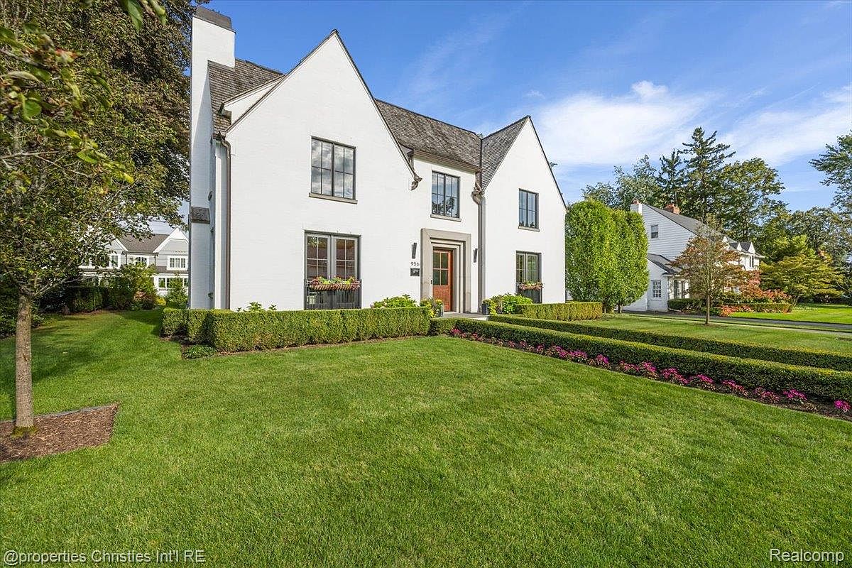 The image showcases the front exterior of a charming two-story house with a well-manicured lawn. The house features a white facade, dark-framed windows, and a symmetrical design with gabled roofs. Lush green hedges and flower beds add to the property's curb appeal, creating an inviting and classic aesthetic.