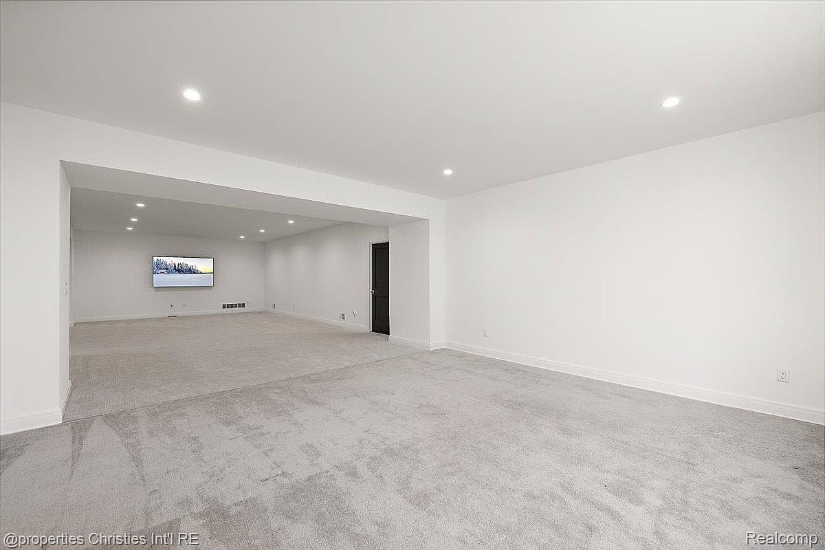 This is a spacious basement area featuring neutral gray carpeting and white walls, creating a bright and open feel. The room is divided into two sections by a large opening, with a television mounted on the wall in the far section. A dark door is visible in the back, and recessed lighting is used throughout the space.