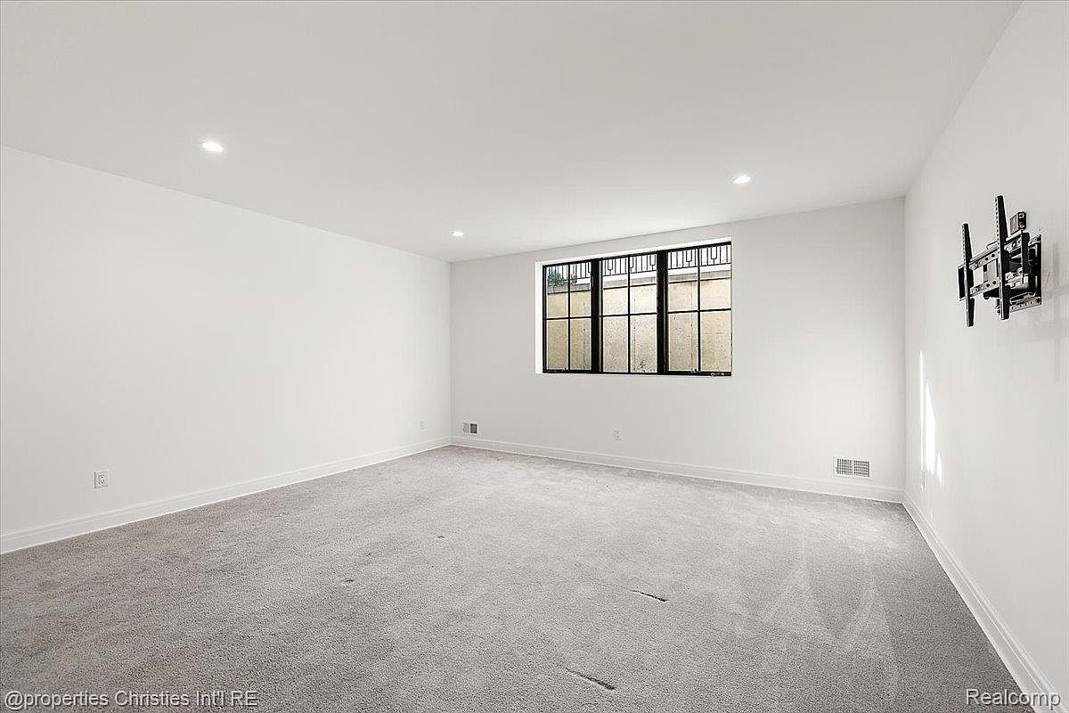 This is a spacious, well-lit basement featuring neutral gray carpeting and white walls. A window with a black frame provides natural light, and recessed lighting enhances the brightness of the room. A wall-mounted TV bracket suggests the potential for a home entertainment area.
