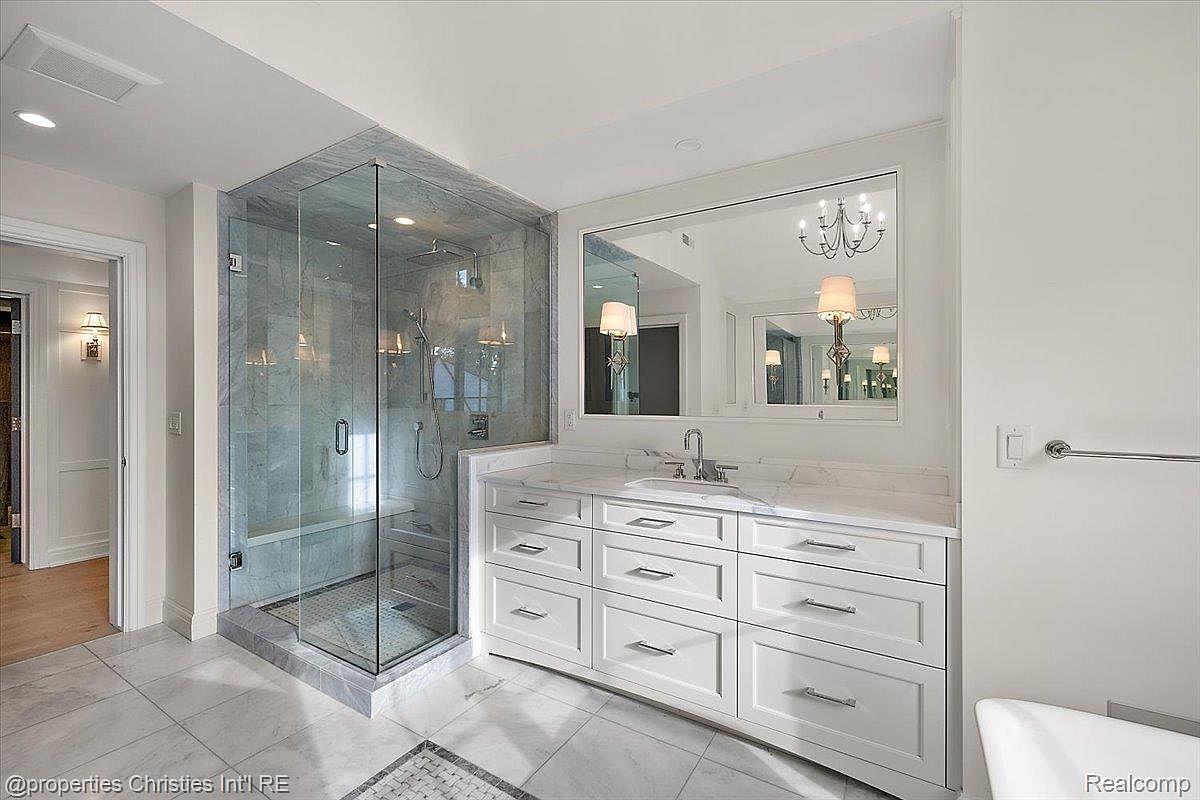 This is a well-lit primary bathroom featuring a glass-enclosed shower with marble-like tiling and a built-in bench. A large vanity with white cabinetry and a marble countertop is complemented by a large mirror reflecting a chandelier, adding a touch of elegance. The flooring is tiled, and the overall aesthetic is clean and luxurious.