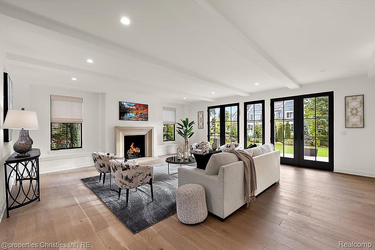 This is a bright and airy living room featuring hardwood floors, white walls, and black-framed windows and doors that offer a view of the outdoors. The room is furnished with a light-colored sofa, patterned armchairs, and a modern fireplace with a television above it. The perspective is from a wide angle, showcasing the spaciousness and natural light of the room.