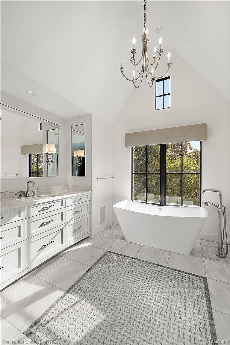 This is a bright and elegant primary bathroom featuring a freestanding white bathtub positioned in front of a large window, allowing natural light to flood the space. The room includes a double vanity with white cabinetry and marble countertops, complemented by a decorative chandelier overhead. The flooring is a mix of large marble tiles and a patterned mosaic rug, adding texture and visual interest.