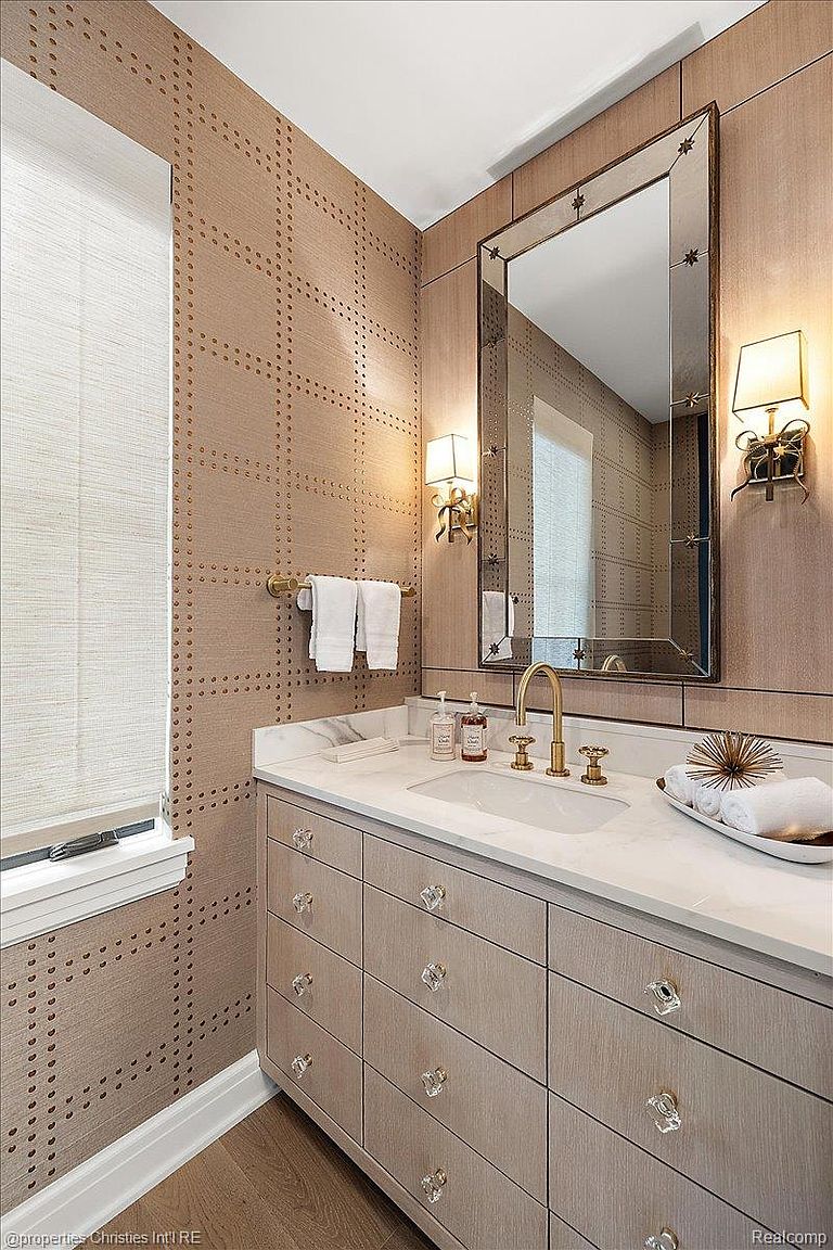 This is a well-appointed guest bathroom featuring a vanity with a marble countertop and crystal knobs. The walls are covered in a textured wallpaper with a grid pattern, complemented by a decorative mirror and sconces. The overall impression is elegant and refined.