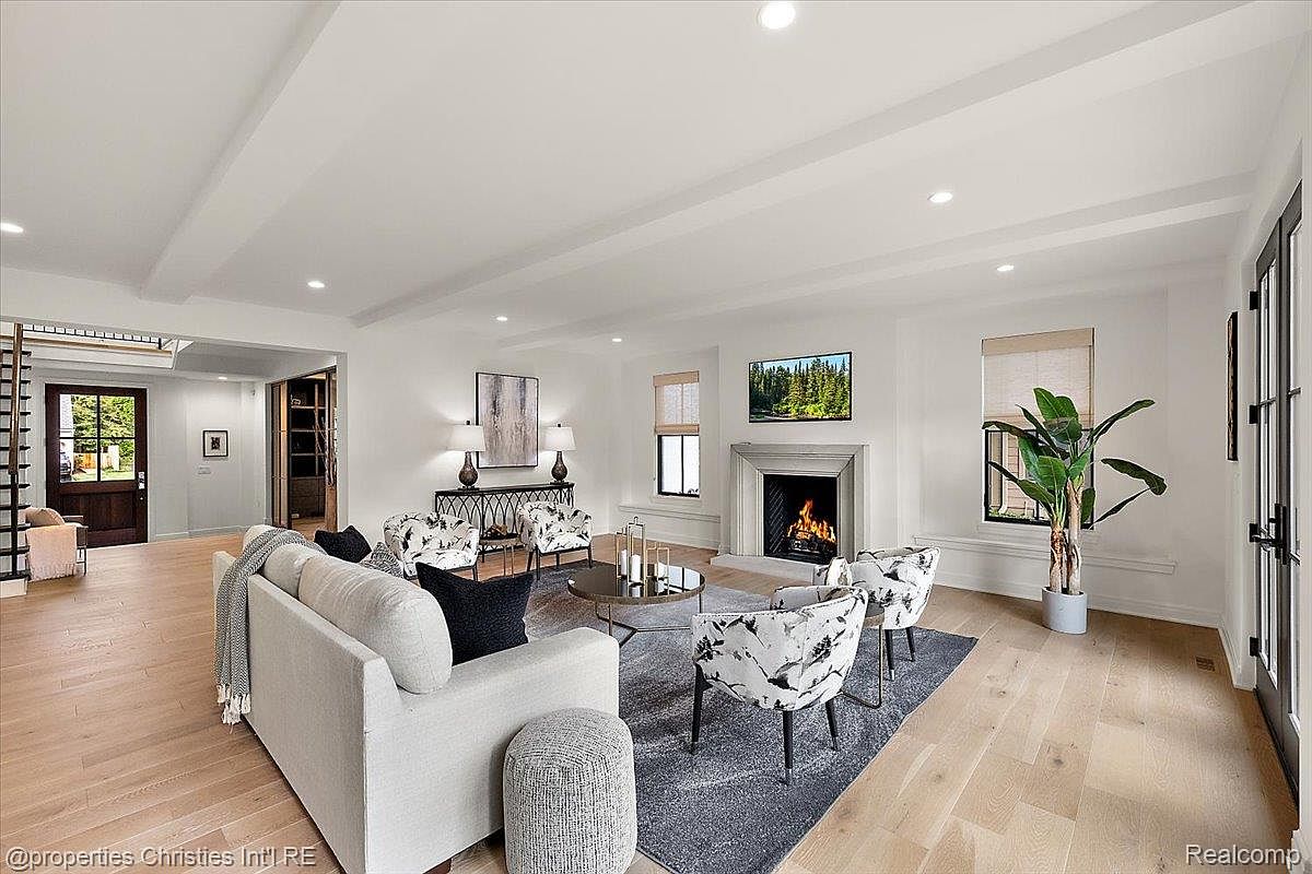 This is a bright and airy living room featuring a neutral color palette with white walls and light wood flooring. The room is furnished with a comfortable sofa, patterned armchairs, and a modern coffee table. A fireplace with a mounted television above it serves as a focal point, and large windows provide natural light, creating a welcoming and sophisticated atmosphere.