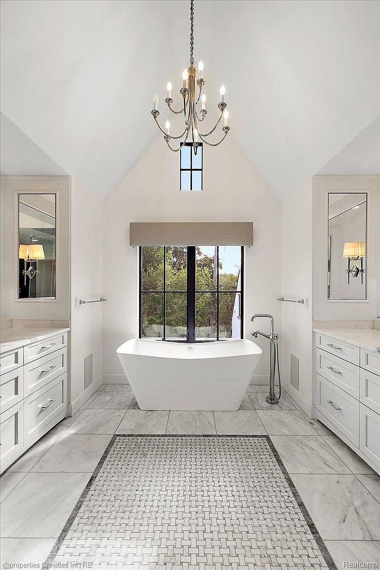 This is a bright and luxurious primary bathroom featuring a freestanding white bathtub centered under a large window, flanked by matching white vanities with marble countertops and framed mirrors. A decorative chandelier hangs above, adding a touch of elegance to the space, which is finished with marble flooring and a patterned tile rug. The perspective is a centered, eye-level shot that emphasizes the symmetry and spaciousness of the bathroom.