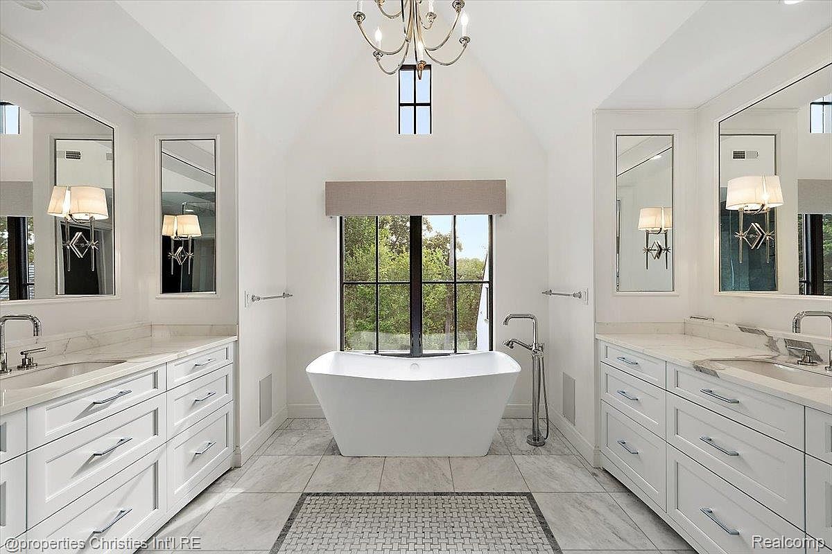 This is a luxurious primary bathroom featuring dual vanities with white cabinetry and marble countertops. A freestanding bathtub is centered beneath a large window, providing natural light. The room is decorated with a chandelier and framed mirrors, creating a bright and elegant space.