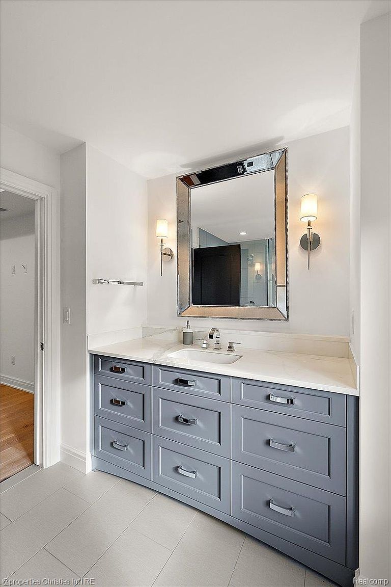 This is a well-lit bathroom featuring a gray vanity with multiple drawers and chrome hardware, topped with a white countertop and a rectangular sink. A large, beveled mirror hangs above the vanity, flanked by sconce lighting. The room has a clean and modern aesthetic, with light-colored tile flooring and white walls.