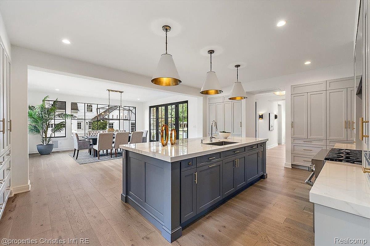 This is a bright and spacious kitchen featuring a large center island with a dark gray base and light countertop, complemented by three pendant lights overhead. The kitchen flows seamlessly into a dining area with a view of the exterior through large windows and doors. The hardwood flooring adds warmth to the space, while the cabinetry provides ample storage.