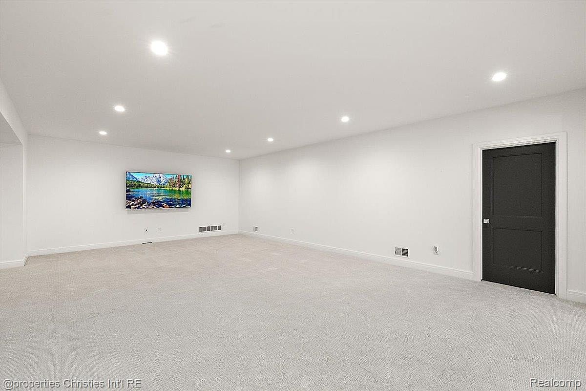 The image showcases a spacious basement area with light gray carpeting and white walls, creating a bright and open feel. A large television is mounted on the wall, and recessed lighting illuminates the room. A dark-colored door is visible on the right side, adding a touch of contrast to the otherwise neutral palette.